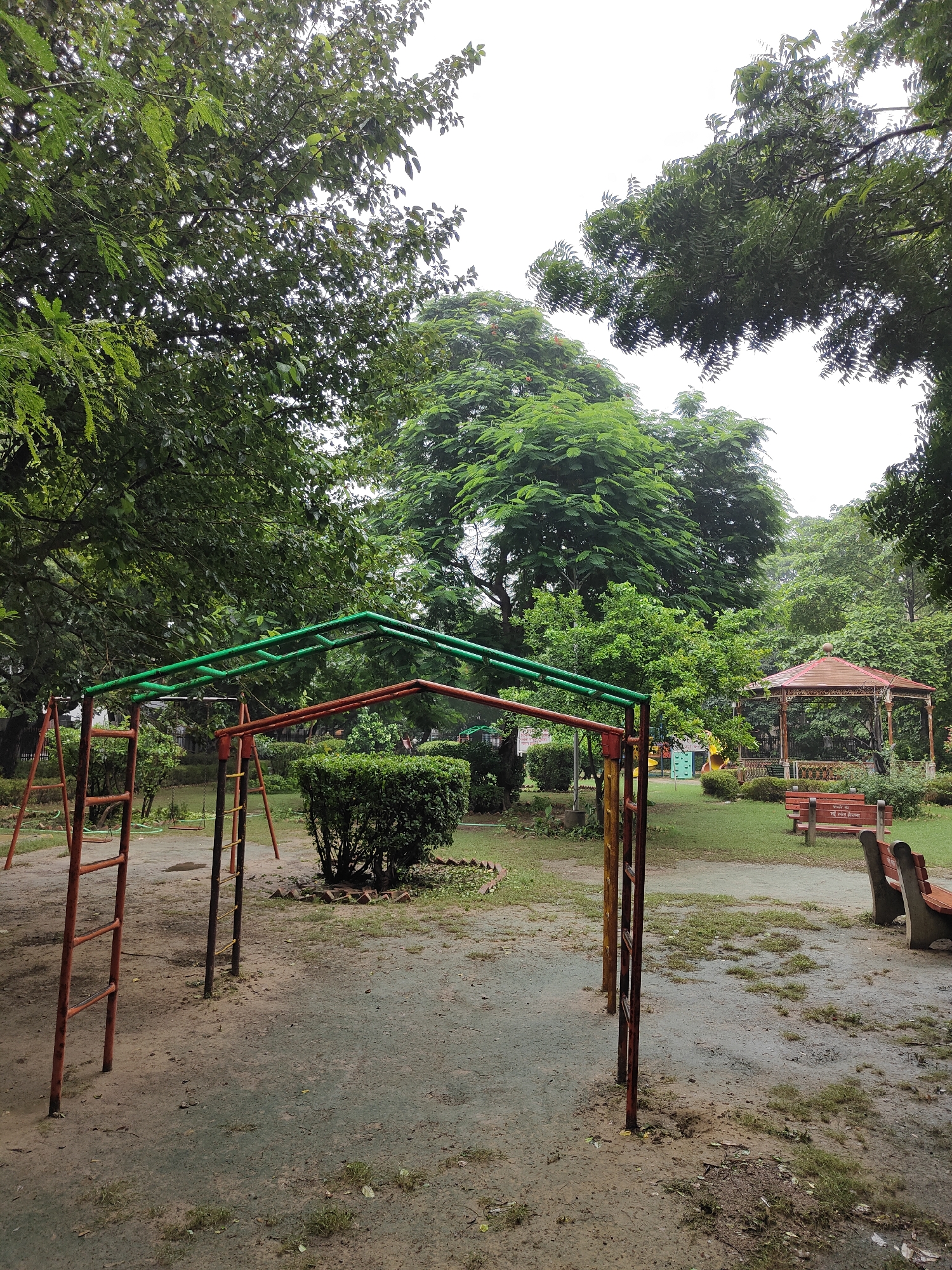 A public park in Gurgaon, India, features a playground with monkey bars and swings. A gazebo stands to the right, with benches in front of it. The park is surrounded by lush green trees, and the ground appears damp, suggesting it may have recently rained. The overall atmosphere is serene and natural.