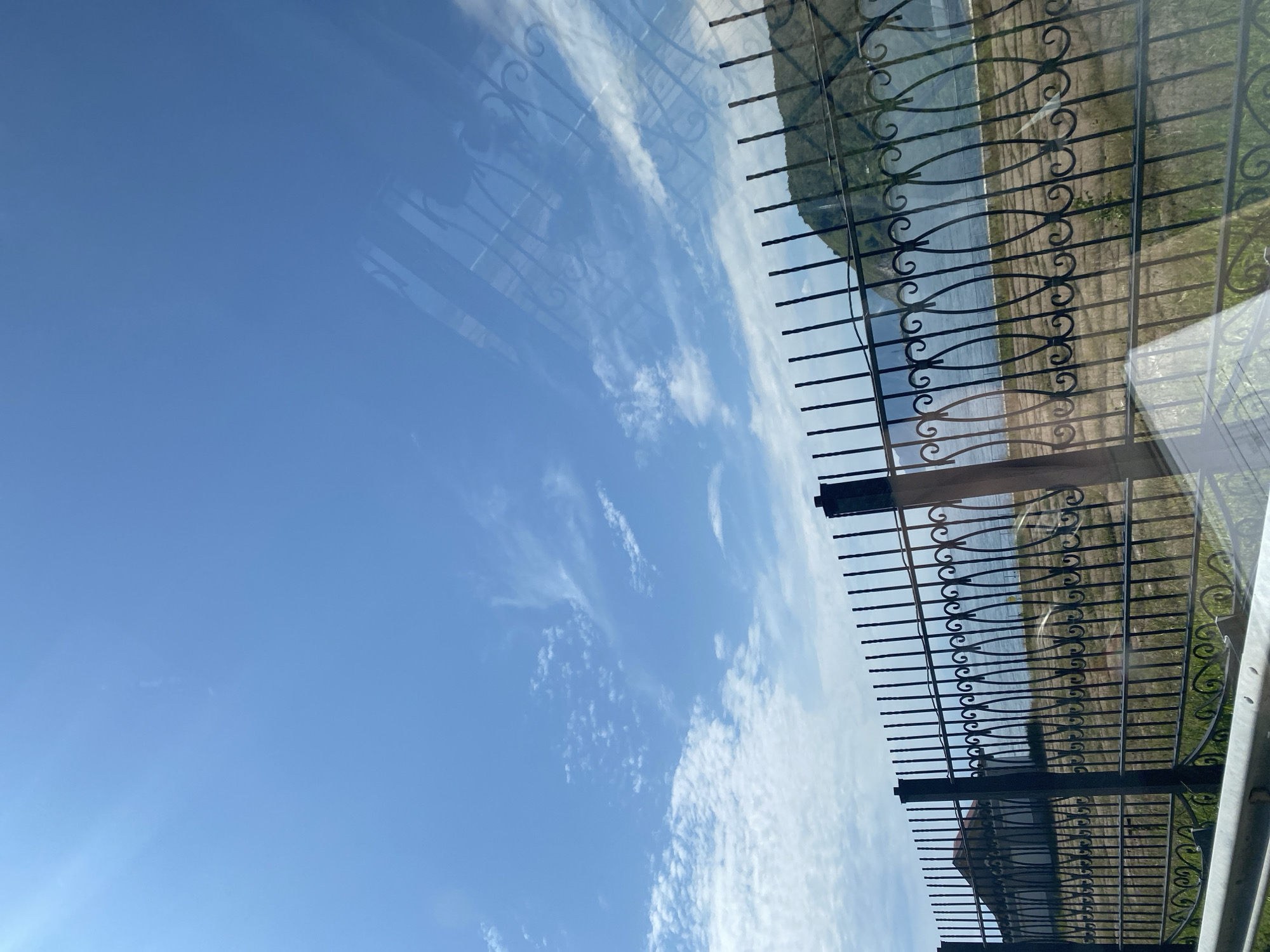 A horizontal, eye-level shot captures a serene outdoor scene with a clear blue sky dotted with wispy clouds. In the foreground, a black wrought-iron fence with decorative scrollwork runs horizontally across the frame, partially obscuring the view of the background. Beyond the fence, a calm body of water, likely Lake Baikal, stretches out, meeting a distant, tree-covered hill under the expansive sky. The grassy ground behind the fence appears dry and sparse, with faint, horizontal lines suggesting cultivated fields. A building with a reddish roof is visible to the far left, peeking over the fence. The overall atmosphere is peaceful and tranquil, characteristic of a summer day in Baikal (Port), Russia.