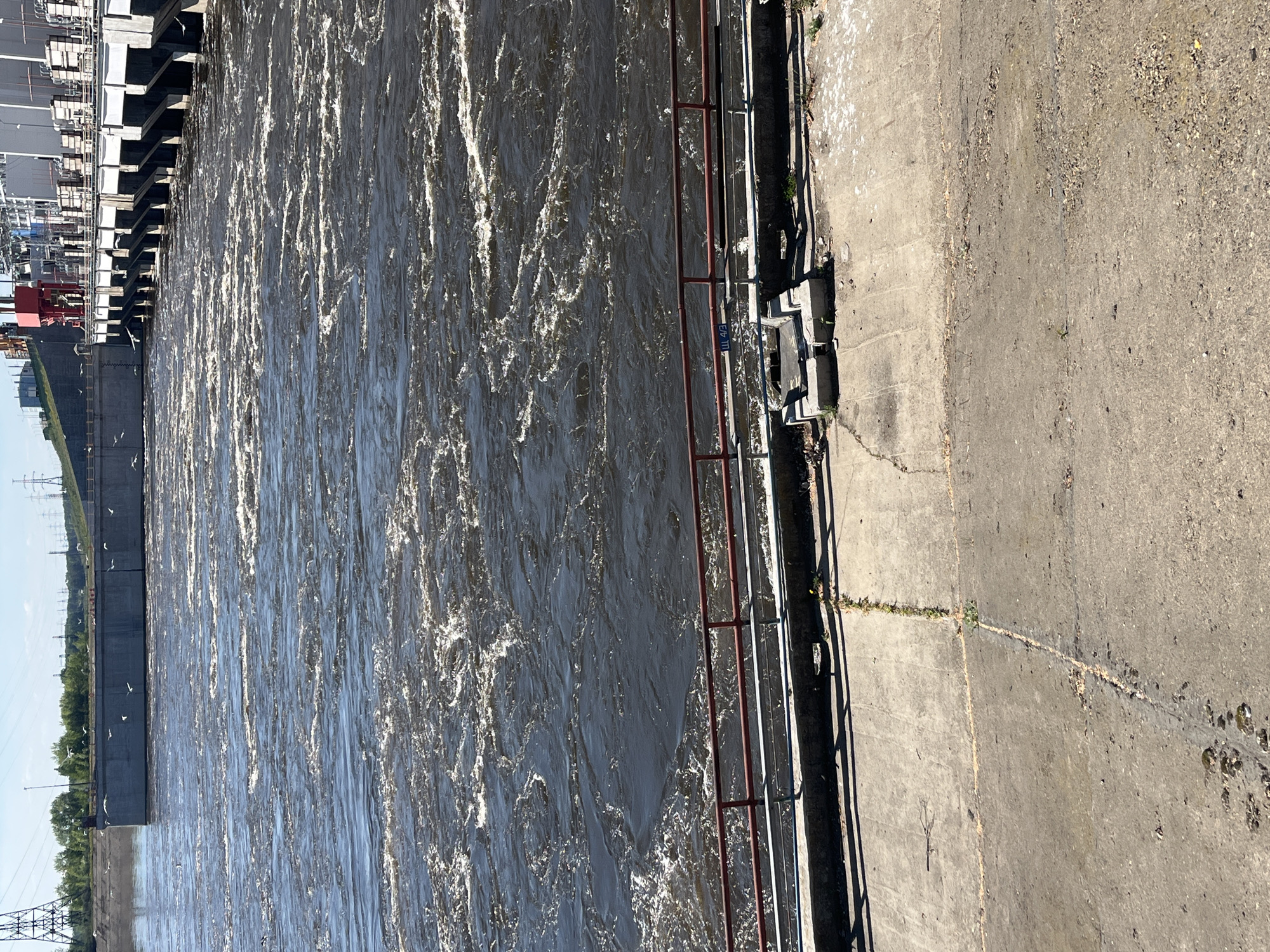A high-angle view shows a turbulent river bordered by a concrete structure with a red and silver guardrail. To the left of the river is a dam structure, visible in the upper left corner, that appears to be part of a power generation facility. Power lines and utility poles are visible in the distance along the left side. On the right, the concrete surface leads to a rougher, gravelly area. Cracks are visible in the concrete, with some vegetation growing within them. The river is dark and churning, reflecting light irregularly. The location, based on the description, is near the city of Noviy, Russia.