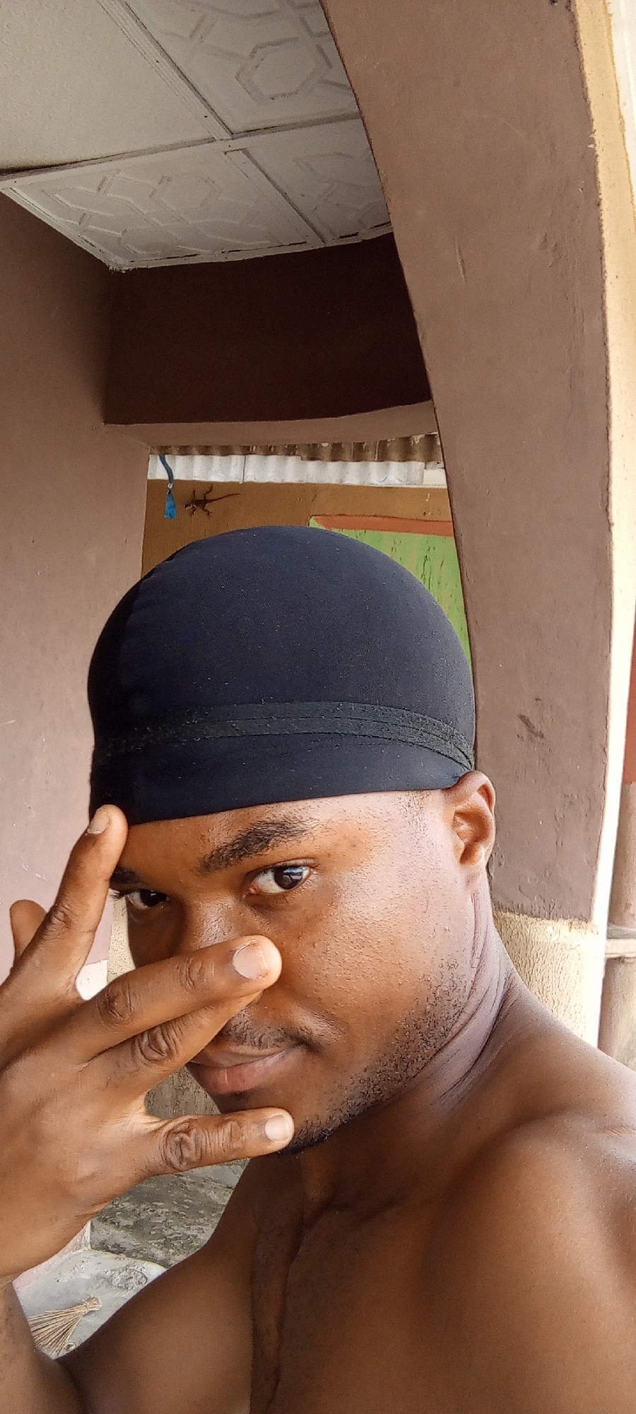 A shirtless, dark-skinned man wearing a black du-rag is in close-up. The man's left hand is raised, with his fingers slightly obscuring his face. The man is partially in front of a painted doorway that has a lizard clinging to the wall. The doorway is situated on an exterior wall with the ceiling protruding into the shot. The location is Ilogbo Eremi, Nigeria.
