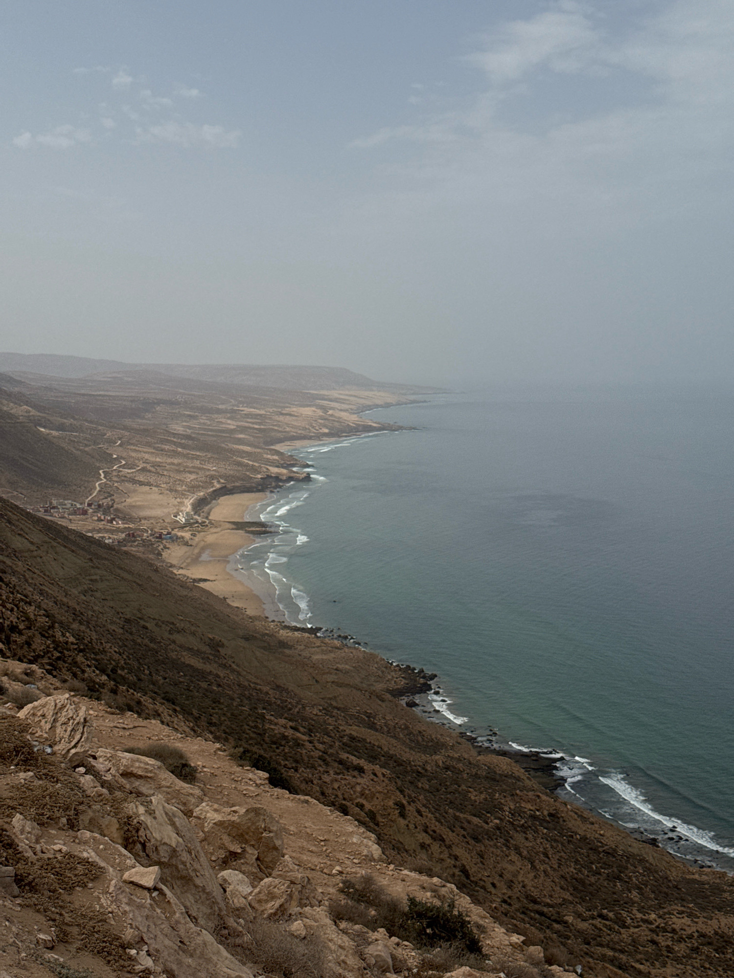 The image shows a coastal landscape near Imsouane, Morocco. A rugged, brown-hued cliff face dominates the foreground, sloping down towards a winding road and a small village nestled on a sandy beach. The village consists of several buildings with red roofs, scattered amidst the arid terrain. Beyond the village, the Atlantic Ocean stretches out to the horizon, its surface a muted teal color, with white waves breaking along the rocky shoreline and the sandy beach. The sky is overcast and hazy, with a few scattered clouds. The overall impression is one of a remote, natural environment with a small human settlement integrated into the landscape.