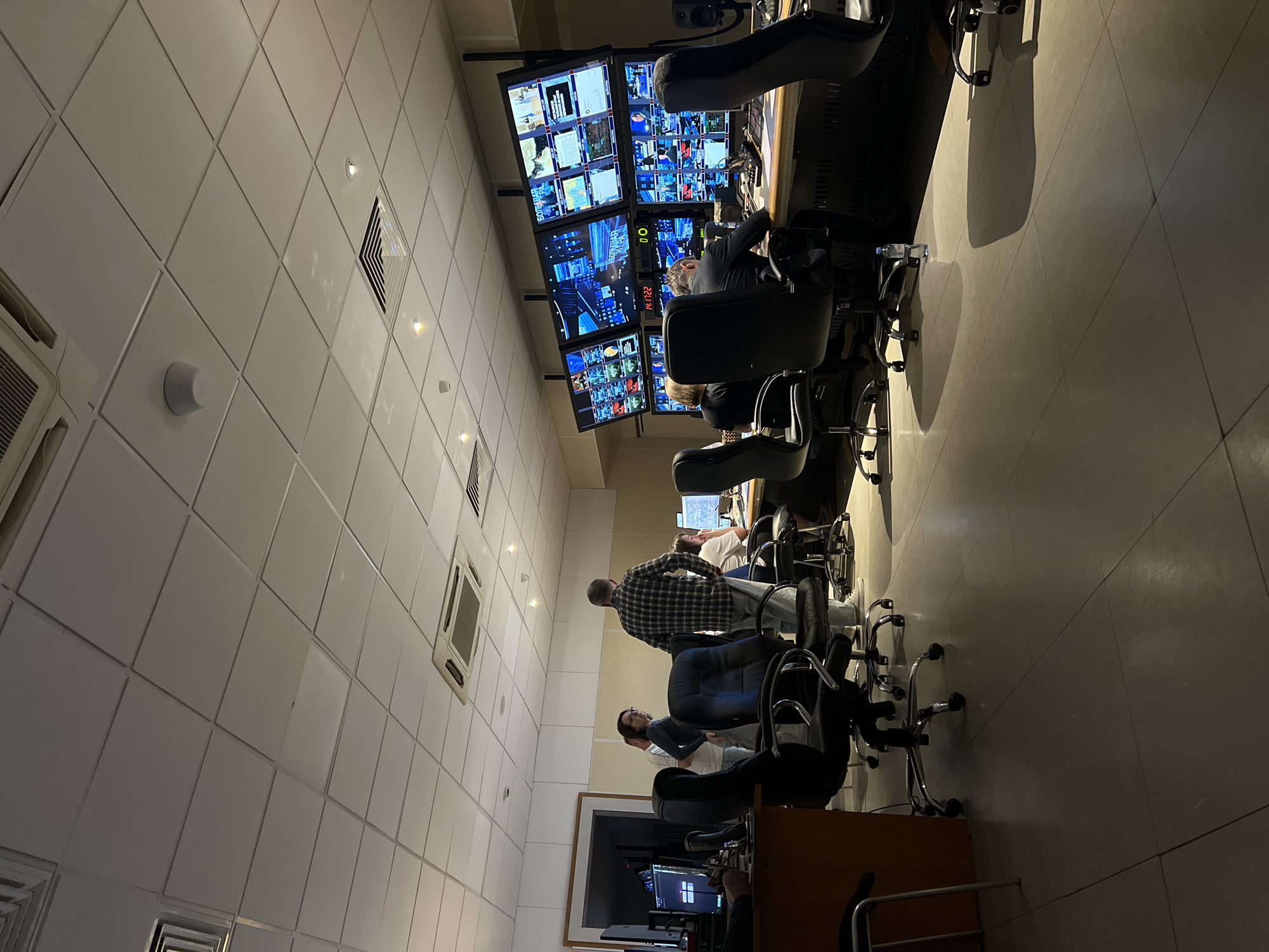 The image shows the interior of a television or video production control room. The room is occupied by several individuals, likely staff, seated in swivel chairs facing a bank of video monitors. These screens display a variety of images, including what appear to be live video feeds, graphic elements, and timecode information. The lighting is a mix of natural light from a window, supplemented with overhead lighting. The walls are tiled. There are air conditioning vents in the wall. The room appears to be located in a modern facility. No specific geographical features of Khimki, Russia, are discernible in the image to positively identify the location. The visible monitors, the room's layout, and the seated individuals indicate a working environment focused on video production and control.