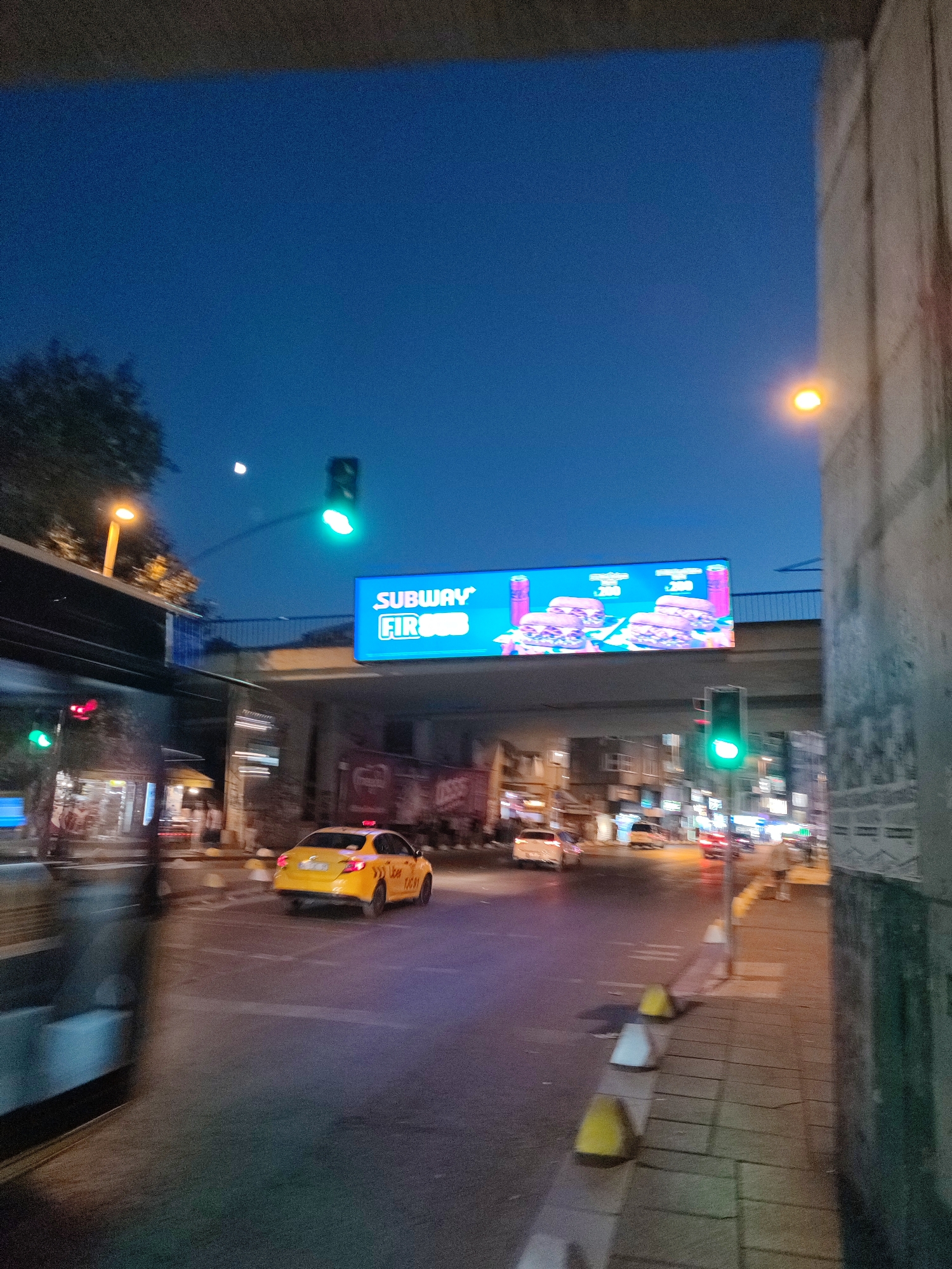 The image depicts a street scene at dusk in Kadıköy, Turkey. A yellow taxi, identified by "Uber" branding and the license plate "34 JU 6091", is driving on the right side of the road. An overhead billboard displays an advertisement for Subway. Traffic lights are visible, with one showing a green signal. The street is illuminated by streetlights and the ambient light from buildings, reflecting on the wet asphalt. A sidewalk with yellow and white traffic cones runs along the right side of the image. The environment suggests a bustling urban area, with some motion blur indicating movement.