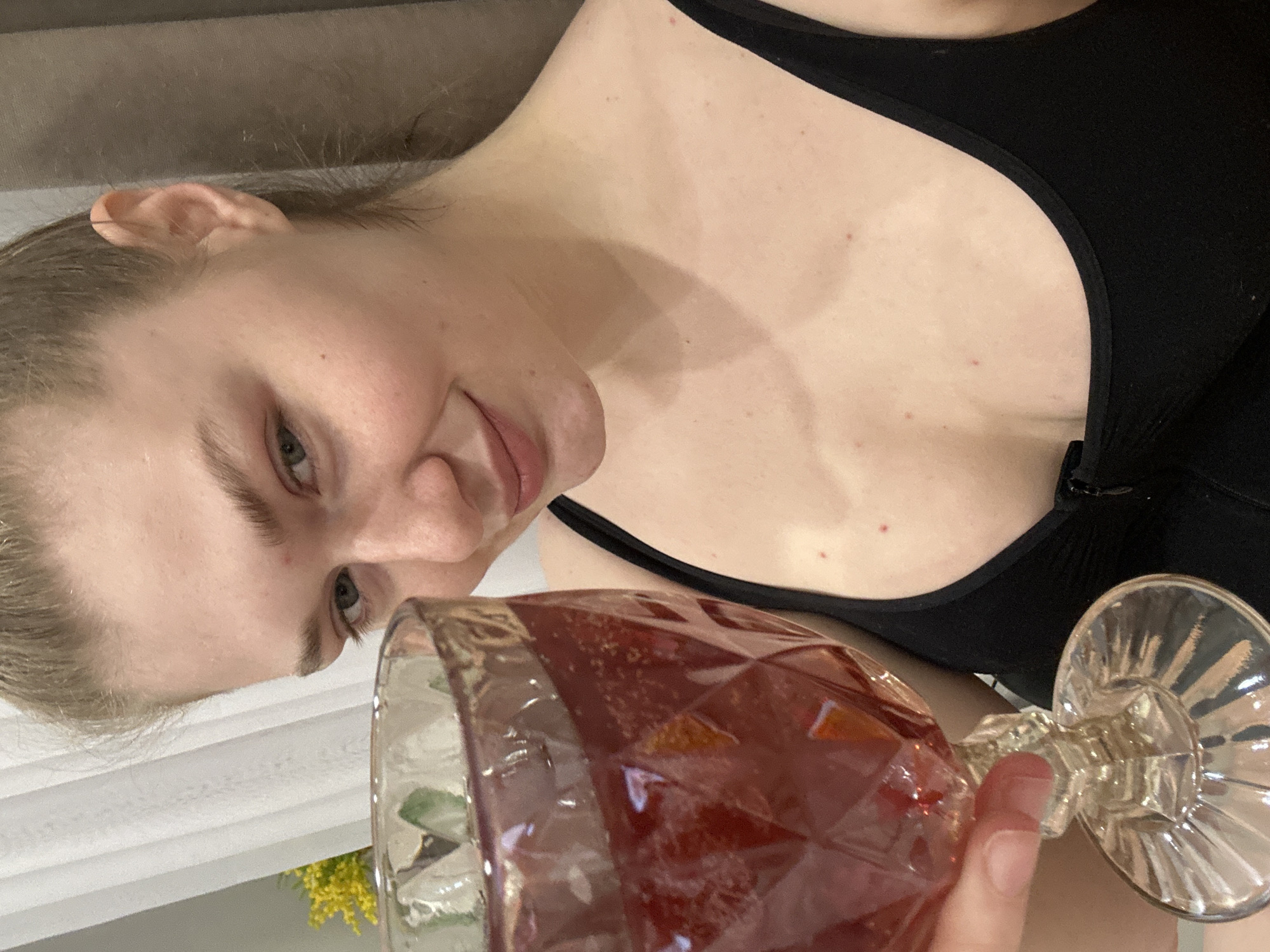 A person with fair skin and blonde hair holds a stemmed glass filled with a red liquid. The person wears a black tank top. Red spots are visible on the chest. A couch is partially visible in the background. Based on other images, the location may be Kazan, Russia, although that information is not explicit in this single image.