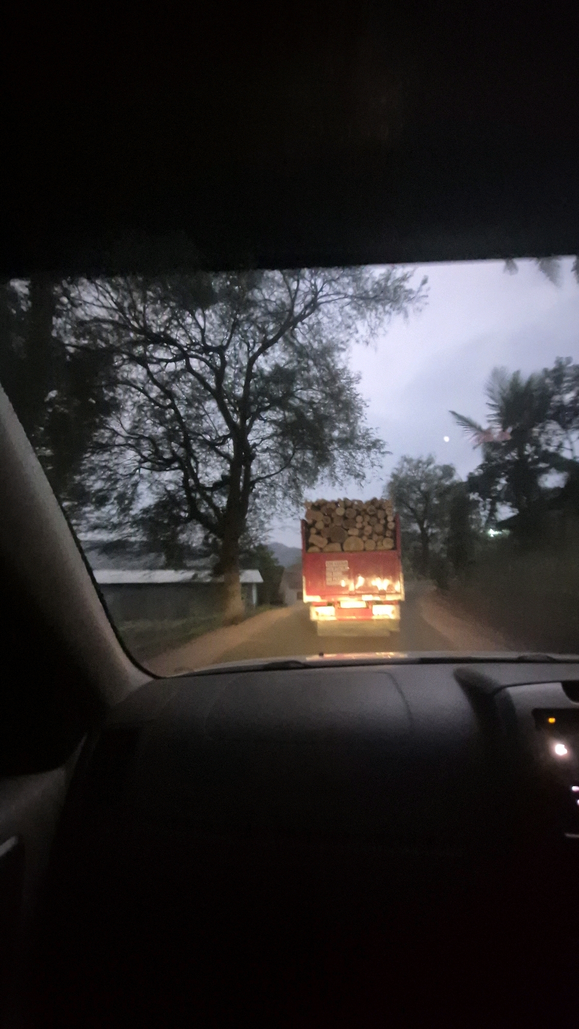 The image shows a view from inside a vehicle, looking forward onto a two-lane road. A red flatbed truck is visible in the distance, carrying a large load of cut logs. The truck's brake lights are illuminated. The road is flanked by trees, some of which are bare of leaves. There are also some buildings visible on the left side of the road. The sky is overcast and the lighting suggests it is either dawn or dusk. The interior of the car is dark, with the dashboard visible in the foreground. The location is identified as Batuwarno, Indonesia.