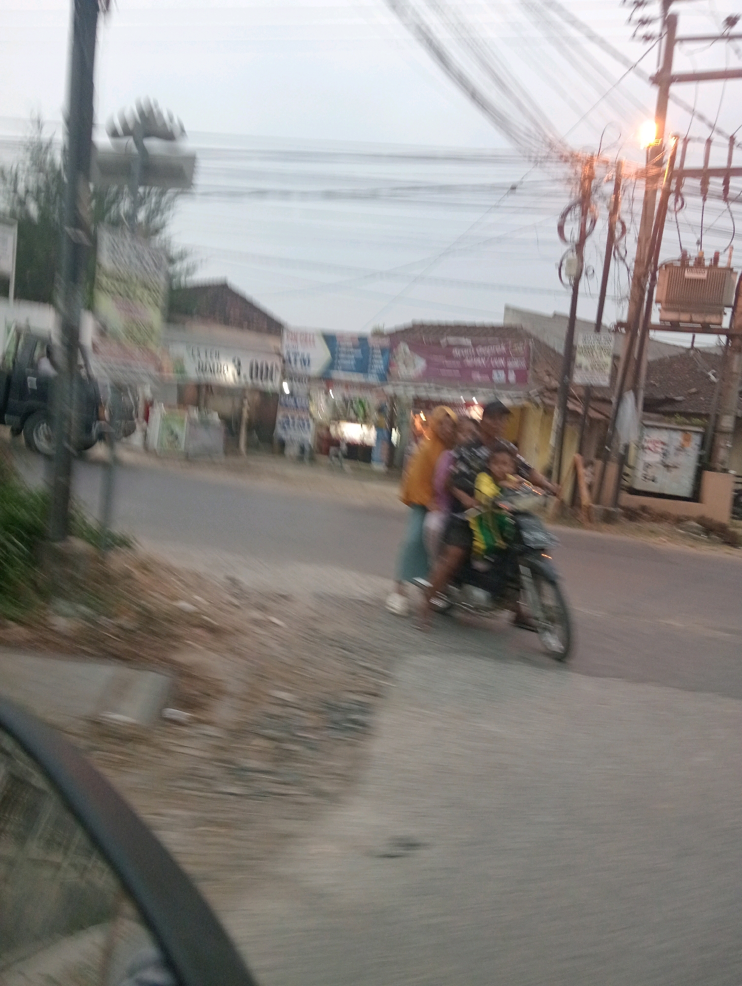 In the city of Pringsewu, Indonesia, the image captures a blurred scene of a motorcycle with three occupants traveling on a street. A person wearing a yellow headscarf and an orange top is seated behind the driver, who is wearing a checkered shirt. A child in a yellow shirt is positioned in front of the driver. The motorcycle is dark in color. In the background, there are buildings with signage, including a prominent sign that appears to say "ATM". Electrical wires and poles are visible overhead, with a transformer mounted on one of the poles. To the left of the road, there is a dark-colored utility vehicle parked, and vegetation along the roadside. The overall image is characterized by motion blur, indicating movement at the time of capture.