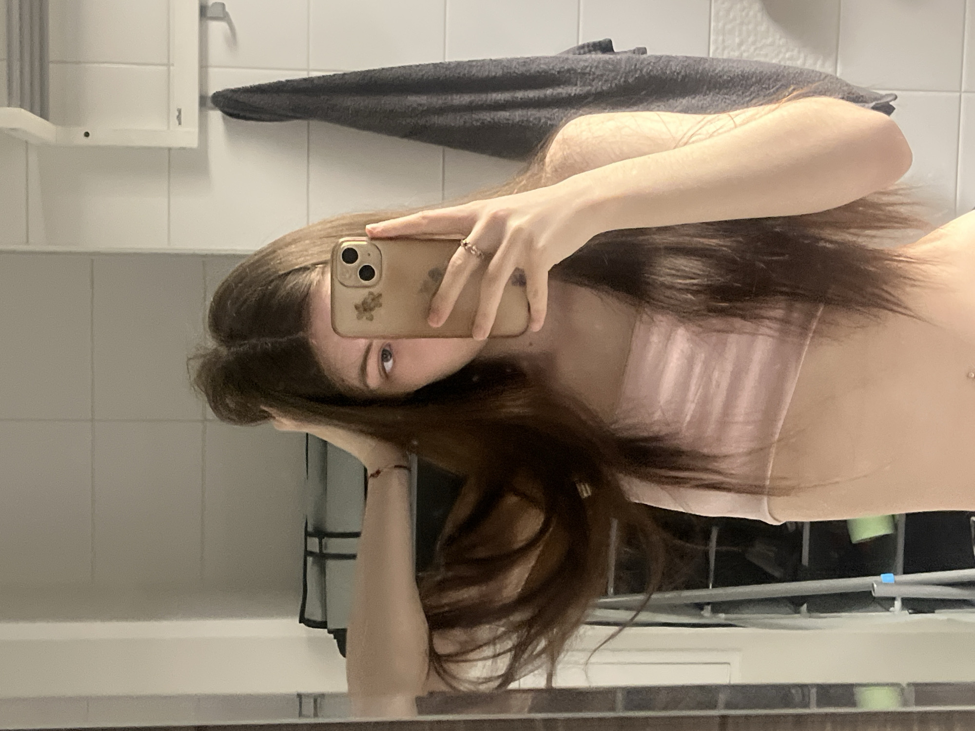 A mirror selfie captures a person appearing female, with long, dark brown hair, in an indoor setting identified as a bathroom in Saint Petersburg, Russia. The individual wears a light pink crop top, exposing the midriff where a navel piercing is visible. Their right hand, adorned with a ring, holds a gold-toned iPhone featuring a three-camera module and a floral design on its case, partially obscuring the face, though the right eye is visible. The left hand is raised, seemingly interacting with the hair near the top of the head. The background consists of white tiled walls. Reflected in the mirror on the right is a dark gray towel hanging on a hook, and a white, slatted wall-mounted shelf above it. On the left side of the reflection, a shelving unit is visible, with a roll of green material resting on one of its shelves.
