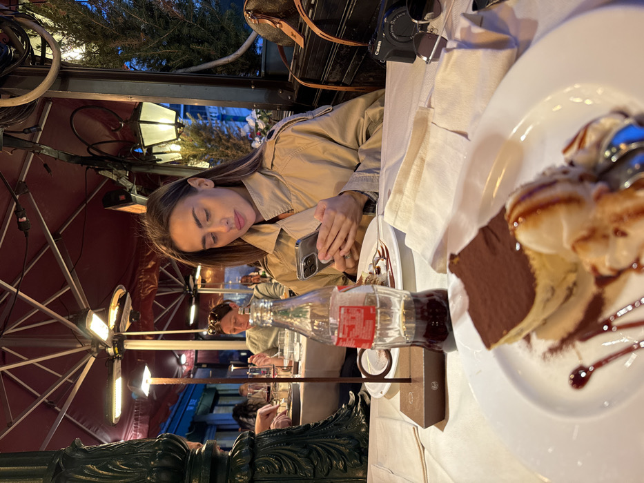 The image captures an evening scene at an outdoor restaurant in Venice, Italy. A woman with long brown hair, wearing a beige trench coat, occupies the midground, seated at a table with a white tablecloth. She is actively engaged with a silver-colored smartphone, holding it with both hands and looking down at its screen. A ring is visible on her left ring finger.
In the foreground, a white plate holds a partially consumed dessert, consisting of a slice of tiramisu topped with cocoa powder and two scoops of vanilla ice cream, garnished with chocolate syrup and a red berry coulis. A half-full glass bottle of Coca-Cola rests in a dark metal stand. To the right, a folded white cloth napkin, a black Canon mirrorless camera with its lens cap on, and dark sunglasses are arranged on the table.
The restaurant's outdoor seating area is covered by large, dark red awnings or umbrellas, from which multiple electric heaters and various light fixtures, including decorative lanterns and smaller table lamps, are suspended, providing illumination. In the blurred background, other patrons are visible at adjacent tables, including a woman with sunglasses perched on her head. Architectural details of the building and some green foliage are discernible behind the main subject. A brown monogrammed bag rests on a ledge to the far right. The overall lighting suggests the time of day is dusk or night.