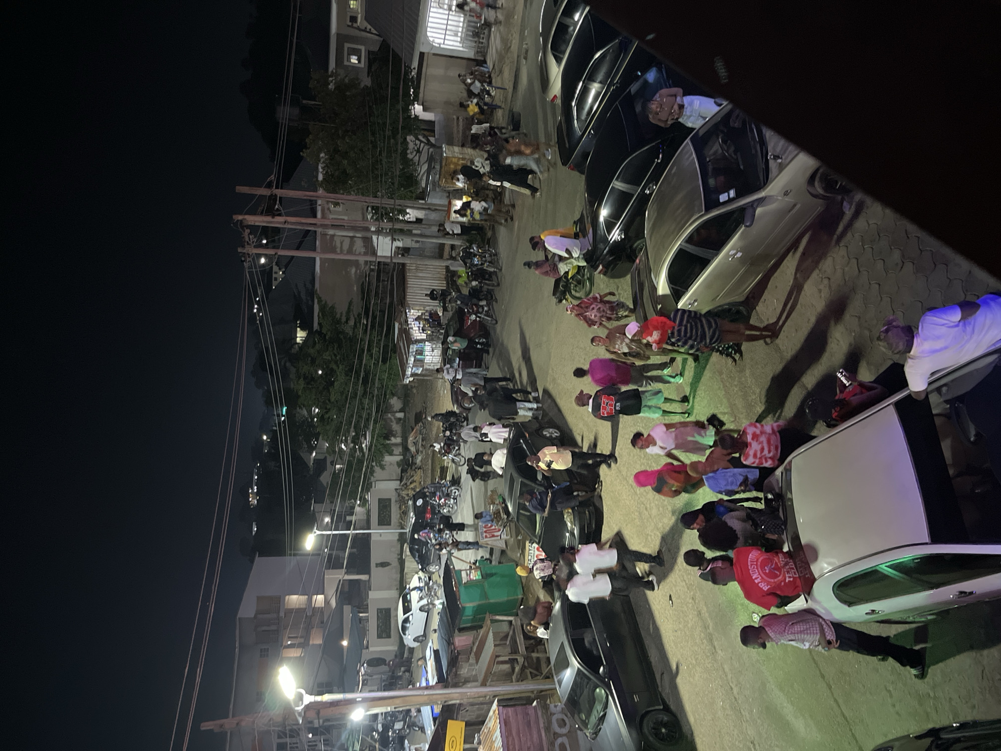 An eye-level, outdoor, nighttime photograph captures a street scene with numerous people and parked vehicles. Several individuals are standing around, some appearing to converse while others are seated or walking. A variety of cars, including sedans and possibly a truck, are parked along the street and on what appears to be a paved lot. Motorcycles are also visible, some parked and others potentially being operated. Several market stalls or small shops are set up along one side of the street, with some appearing open and others closed. Overhead power lines and streetlights illuminate the scene. The background reveals buildings and trees, indicating an urban or suburban environment. The overall impression is one of a lively gathering or marketplace after dark.