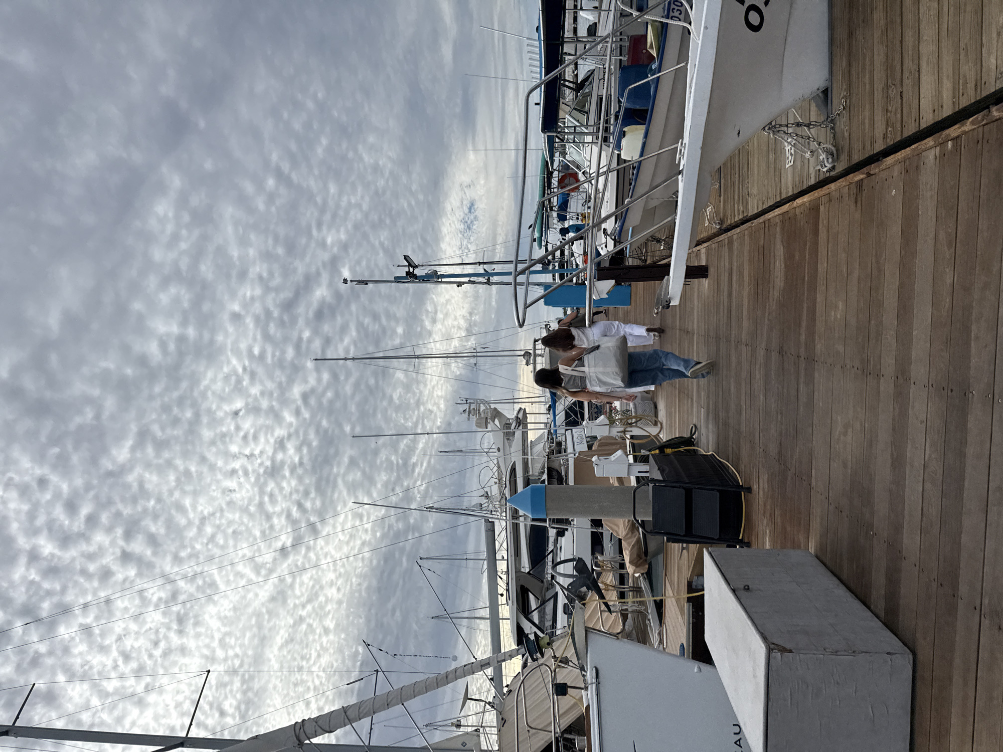The image depicts a marina setting in La Paz, Mexico. Several sailboats and yachts are moored alongside a wooden pier. Two individuals, one in a white top and the other in a light-colored top and jeans, are walking along the pier between the boats. The sky is overcast with textured cloud cover. A black rolling cart is present on the pier near one of the boats.