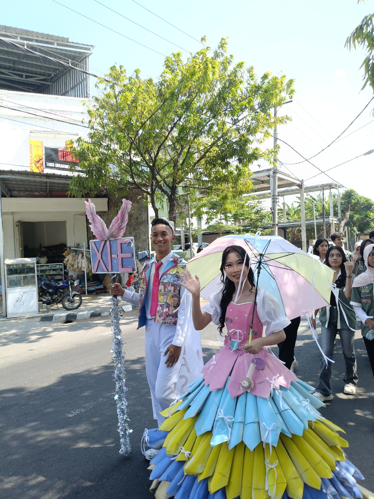 In the city of Kediri, Indonesia, on a sunny day, a young man and woman are participating in a parade. The man, dressed in a white shirt, pink tie, and a colorful vest adorned with various printed images, holds a tall, silver-tasseled staff topped with large pink feather-like decorations and a sign with the letters "XE". The woman, wearing an elaborate dress made of folded pastel-colored fabric resembling petals, walks beside him, holding a colorful umbrella. Her dress is primarily pink at the top, transitioning to blue and yellow at the bottom. Several other young people are visible in the background, some holding bottles and looking towards the camera. The parade appears to be taking place on a paved street lined with buildings and trees. In the foreground, a motorcycle is parked on the side of the road. The overall scene depicts a festive outdoor event.