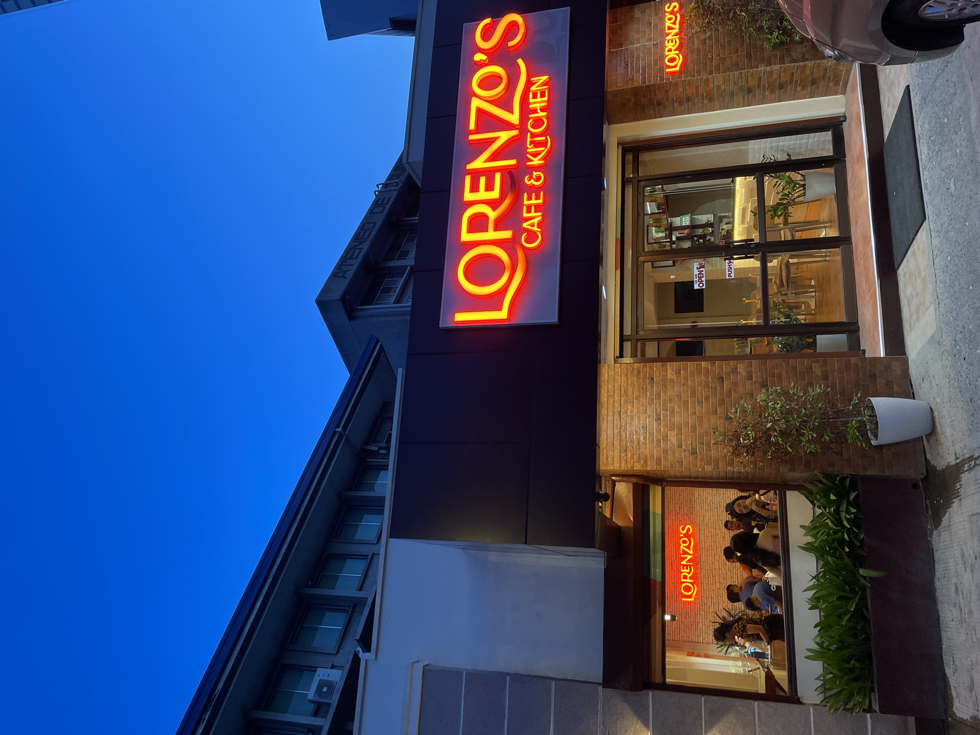 The image depicts the exterior of "Lorenzo's Cafe & Kitchen" located in Davao City, Philippines, at dusk. The cafe's facade features a prominent neon sign in bright orange, spelling out "LORENZO'S CAFE & KITCHEN". To the left, a large window offers a view of the cafe's interior, where several patrons are seated at tables. The building has a modern architectural style with brick and dark paneling elements. Above the cafe, a multi-story building with numerous windows and an air conditioning unit is visible against a deep blue sky. A plant in a white pot stands to the right of the cafe entrance, and part of a car is visible in the lower right corner. The scene suggests a lively evening ambiance at the establishment.