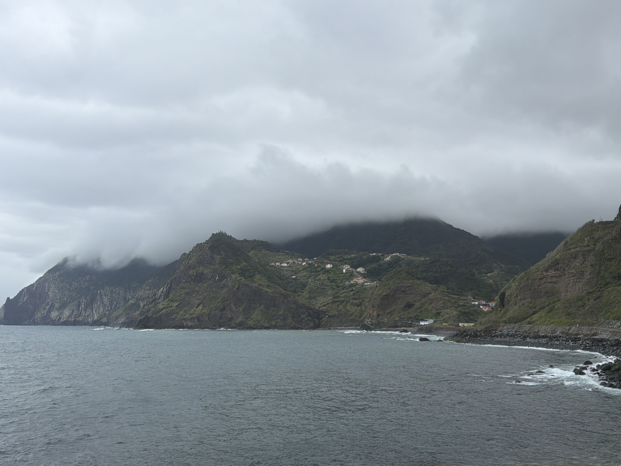 The image shows a coastal scene in Machico, Portugal on a cloudy day. Dark grey water fills the foreground with small waves. Rocky, green cliffs form the coastline. A small number of white buildings are visible on the hillside. Heavy grey clouds obscure the tops of the mountains. A paved road runs along the coastline. The rocks on the shoreline are dark.