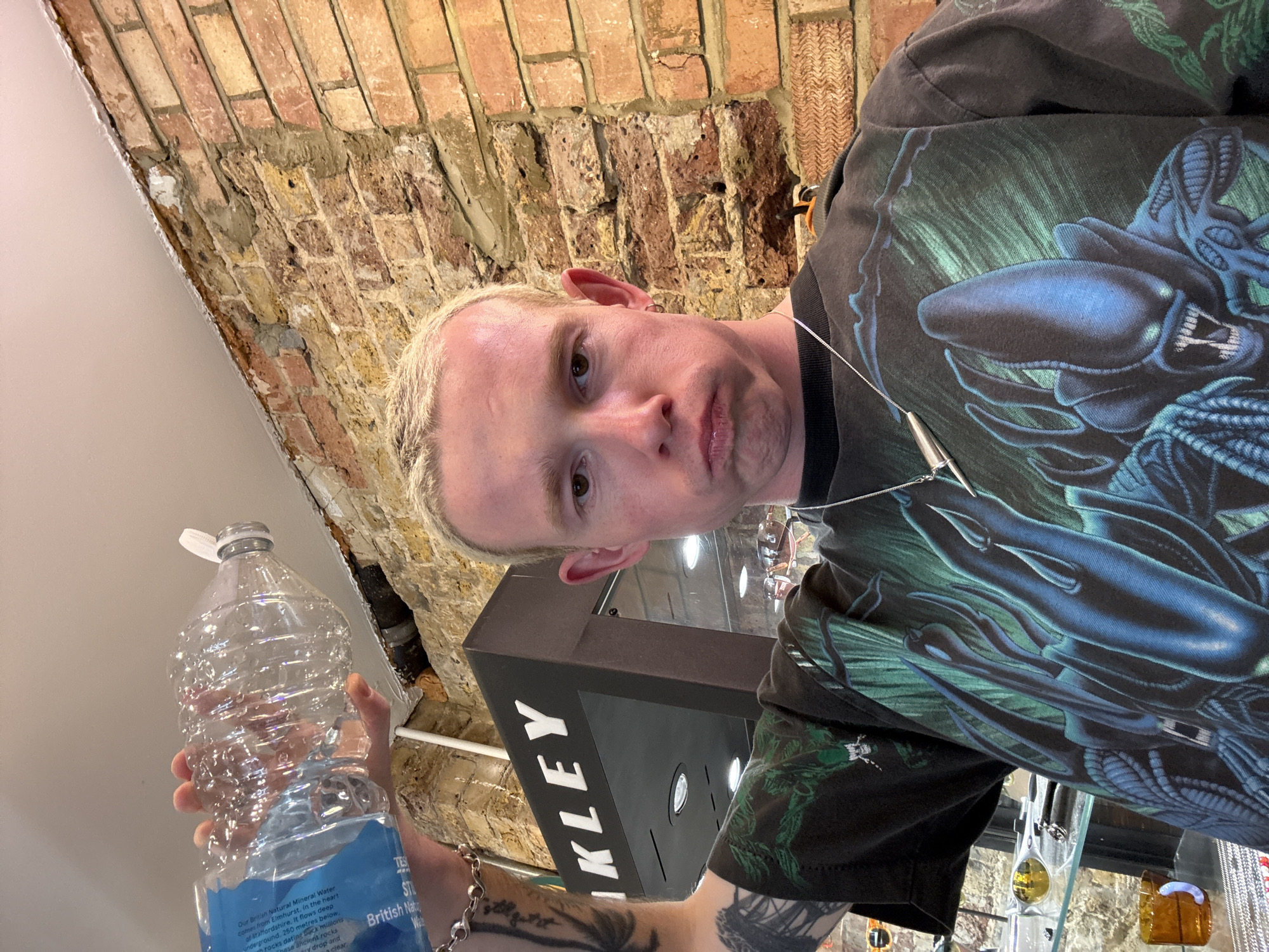 A selfie shows a man with blond hair and brown eyes, wearing a black t-shirt featuring a blue and green alien design, and a silver necklace. He's holding a large plastic bottle of "British Natural Mineral Water" with blue label. The background shows a brick wall and a shop display with "Oakley" branding. The location appears to be a store in London, United Kingdom, based on the mineral water labeling. The man is likely a customer or an employee, as he is standing within the retail environment.