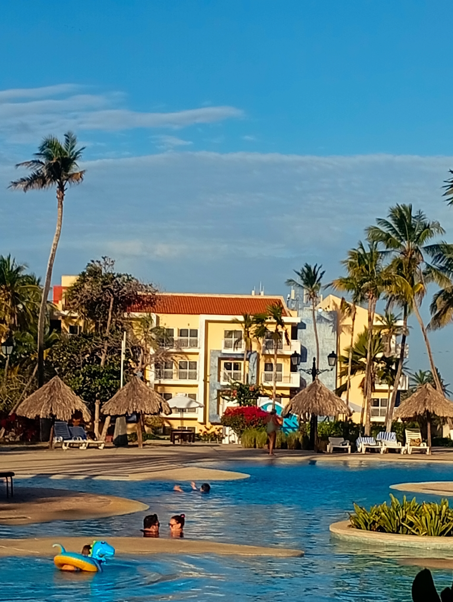 The image depicts a daytime scene at a resort-style swimming pool in Playa El Agua, Venezuela. Several individuals are present in the pool, including two people whose heads are visible above the water's surface near a floating inflatable dinosaur toy. Further back in the pool, there are two more individuals wading. In the background, there is a yellow building with a red roof, partially obscured by palm trees. Surrounding the pool are thatched-roof umbrellas and lounge chairs. The sky is blue with scattered clouds.