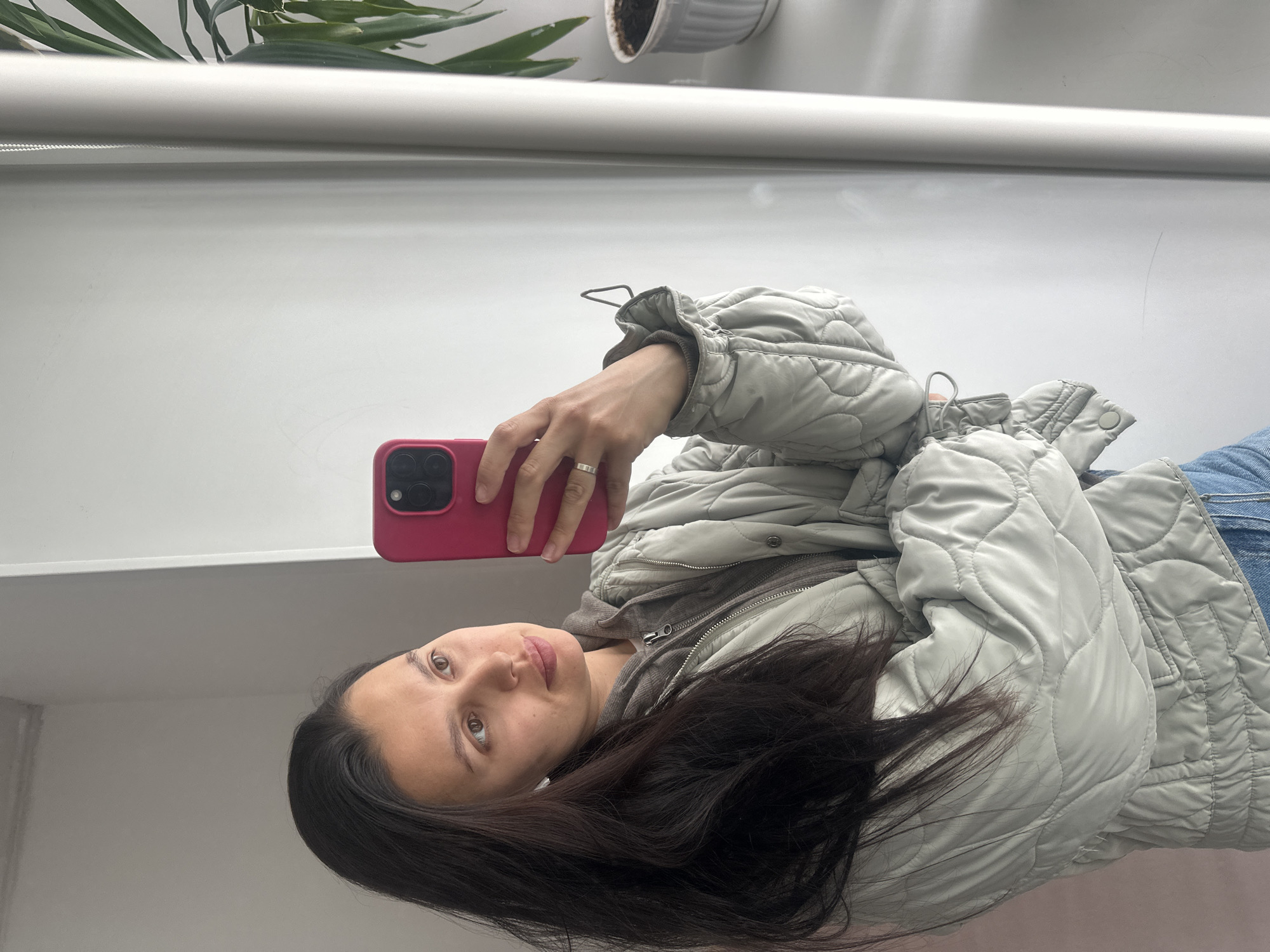 A woman with long dark hair is taking a selfie with a pink phone. She is wearing a light grey quilted jacket and blue jeans. A hooded sweatshirt is visible underneath the jacket. A plant in a white pot sits on a windowsill behind her. The background is a white wall. The location is not discernible from the provided image. There is no definitive evidence to suggest this image was taken in Chaikovsky, Russia.