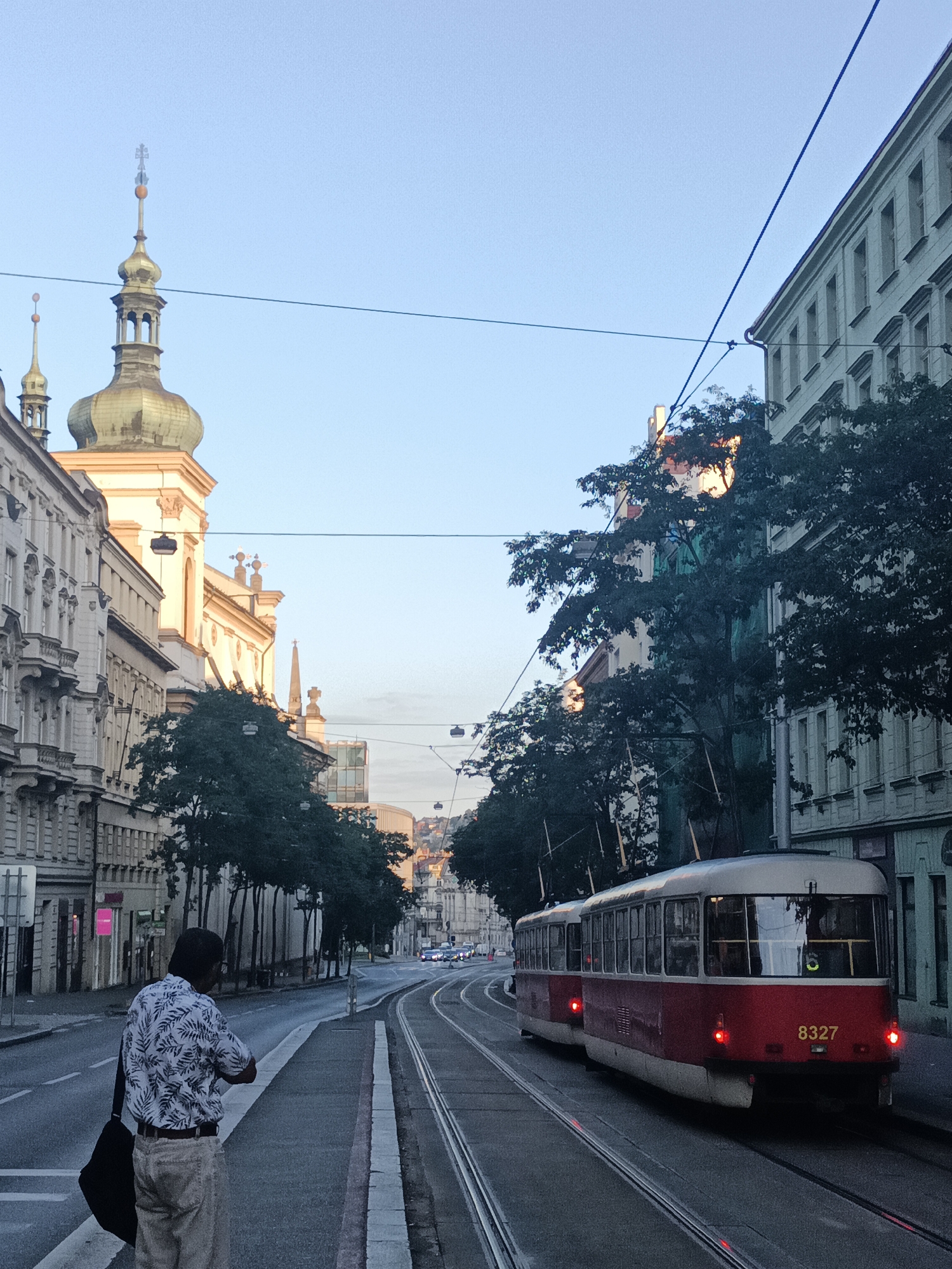 An outdoor, eye-level shot captures a street scene in Prague, Czechia, under a clear, pale blue sky at dawn or dusk. The perspective is from a sidewalk looking down a wide street with tram tracks. On the left, a man with dark hair, wearing a white short-sleeved shirt with a dark leaf pattern and light-colored pants, stands with his back to the viewer, carrying a black bag over his right shoulder. He is facing the street and appears to be looking at something.
The architecture lining the street is characteristic of European cities, with ornate, multi-story buildings featuring balconies and detailed facades. Prominently on the left, a church or historical building with a green, onion-shaped dome and a tall spire is visible, bathed in warm light. Trees with dark green foliage are planted along the sidewalk.
In the middle of the street, two red and white trams are on the tracks. The lead tram, numbered "8327," has its headlights on, and red lights are visible on the rear of both trams. The tram tracks curve to the right, leading into the distance where more buildings and distant hills are visible, along with other vehicles. Overhead, power lines for the trams crisscross the sky. The overall impression is one of a tranquil urban morning or evening.