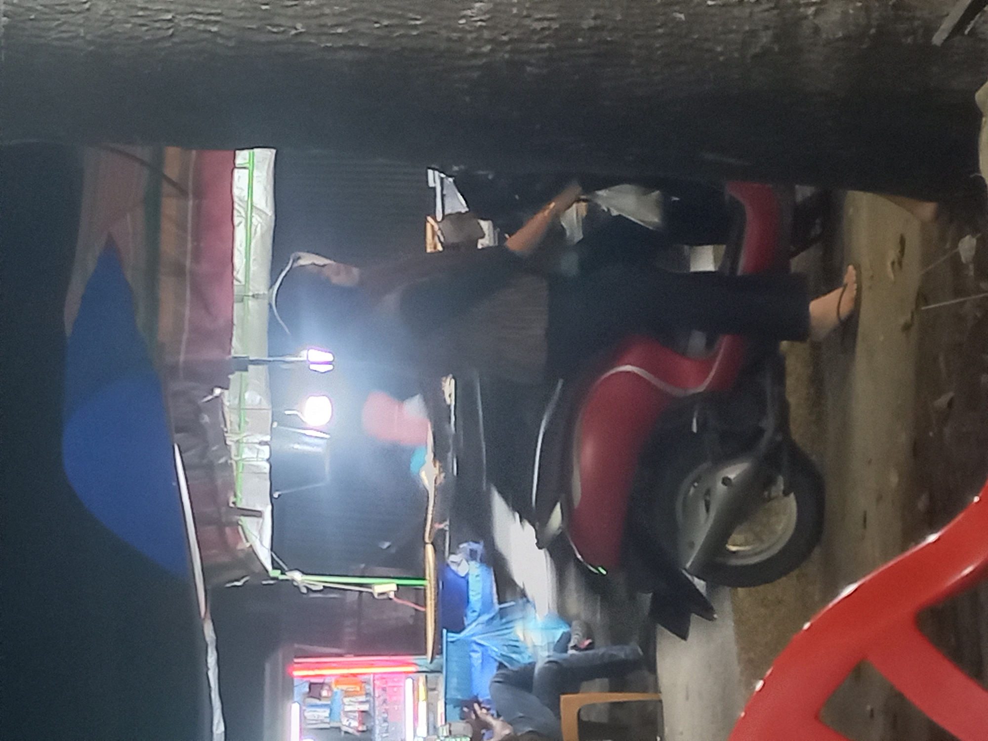 This night-time image, taken in Idi Rayeuk, Indonesia, captures a street scene, likely a market or roadside stall, illuminated by artificial lights.
In the center, a person is seated on a red scooter or motorbike. They are dressed in dark clothing, including black pants, and wear sandals, with one foot resting on a concrete block or a component of the scooter. Their arm is extended forward, suggesting they might be operating or preparing to operate the vehicle, or simply resting.
Behind the scooter, a makeshift stall structure is visible, characterized by overhead coverings of colorful tarps or fabric in shades of pink, light green, yellow, and white, some with a distinct green stripe. Several bright artificial lights illuminate the area, creating noticeable lens flares. A large blue umbrella is open on the left side, partially obscured by darkness, potentially offering shelter from the elements or part of the stall's setup.
Further into the background, another person is visible, seated on a chair and seemingly engaged with a mobile device. Below the stall coverings, there appears to be merchandise or items displayed on shelves, partly obscured by blue plastic wrapping. A red plastic chair is partially visible in the immediate foreground on the right.
The scene conveys a casual, active atmosphere typical of a late-night street or market environment. No discernible text is visible in the image.