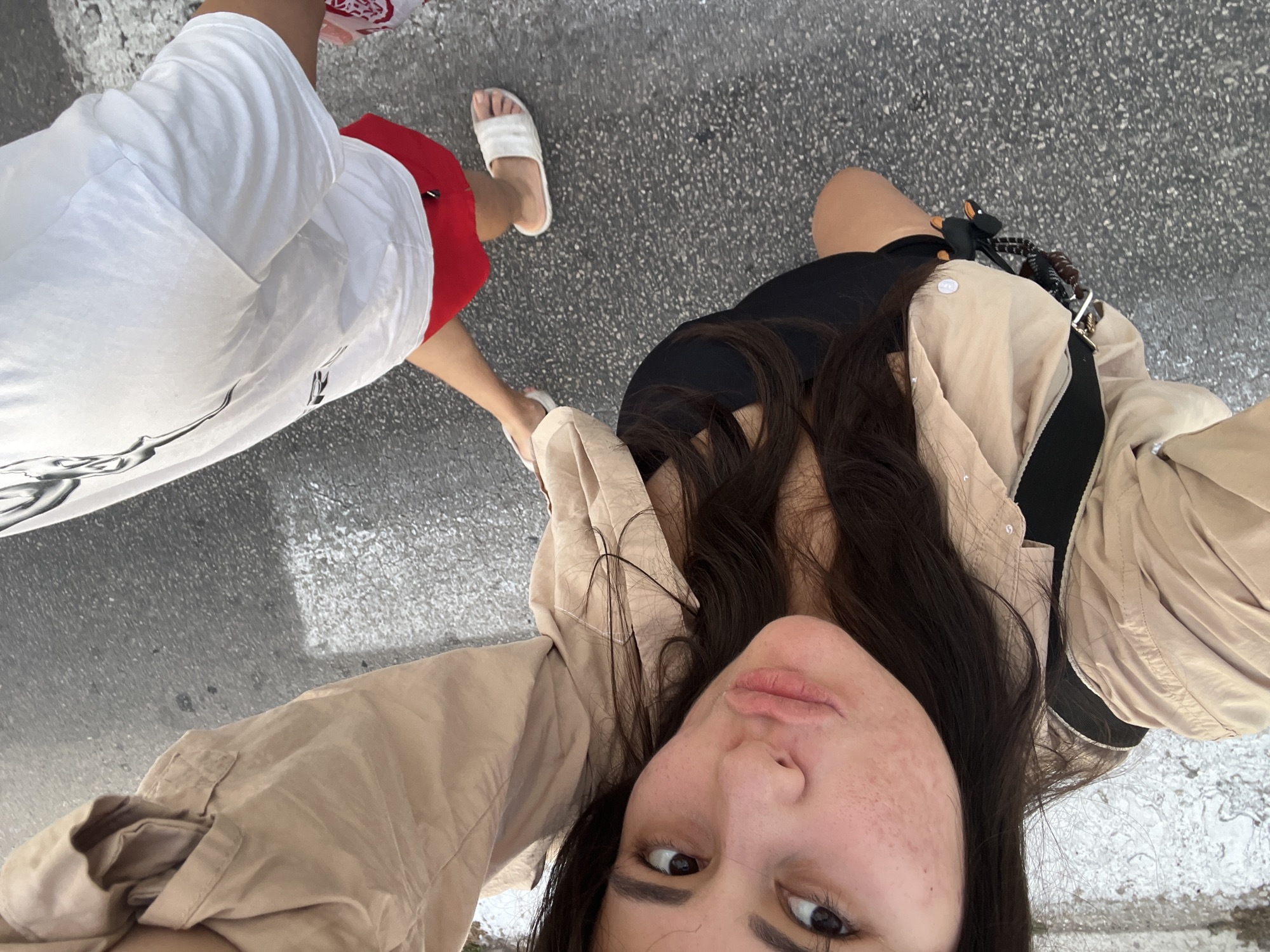 This media file shows a first-person perspective selfie of a young woman with long, dark hair, freckles, and brown eyes. She is wearing a light brown jacket over a black top. Her mouth is slightly open, and her expression could be interpreted as playful or curious. In the background, to her left, are the legs of another person wearing a white t-shirt and red shorts, with white sandals on their feet. The ground beneath them appears to be asphalt.
The image seems to have been taken outdoors during the daytime. The lighting suggests it is sunny. The scene is urban or semi-urban, given the asphalt ground. There are no obvious landmarks to pinpoint the exact location within Kiryat Bialik, Israel, beyond the presence of people in casual attire. No visible text is present in the image. The overall mood is casual and appears to capture a moment of shared activity, possibly walking or posing for a picture, between the two individuals.