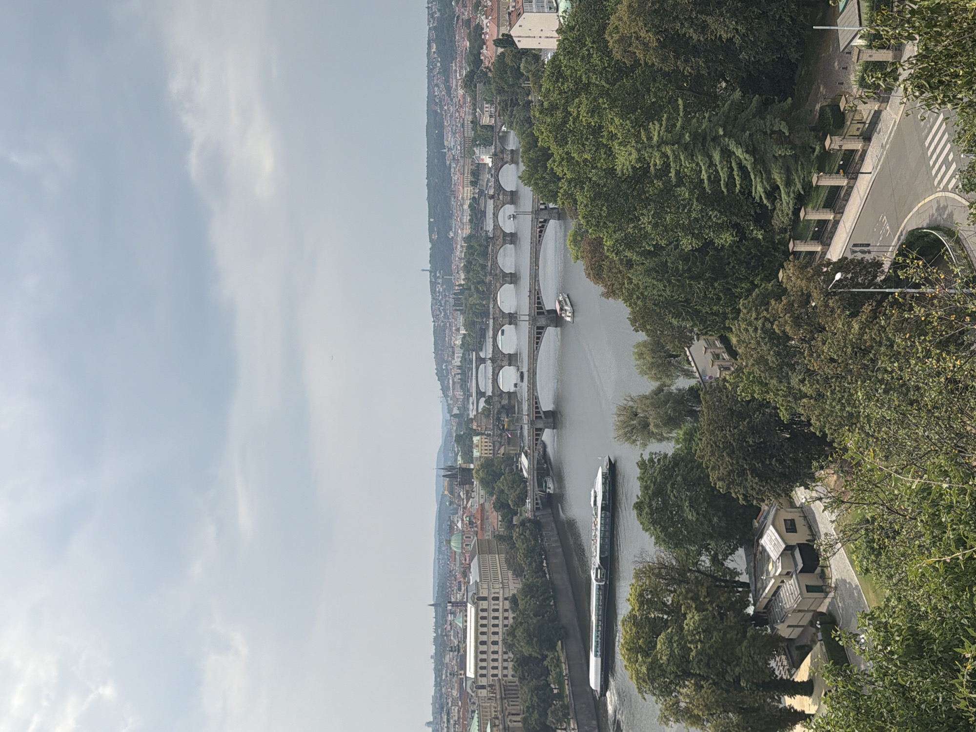 This is an aerial view of Prague, Czechia, showing a wide river with a large tour boat navigating its waters. Multiple bridges span the river, with a city skyline of buildings and church spires visible in the background. Lush green trees and foliage occupy the foreground and sides of the image, with some buildings and a road visible among them. The sky is overcast, with a soft light filtering through the clouds. The scene depicts a scenic urban landscape with a focus on the river and bridges.