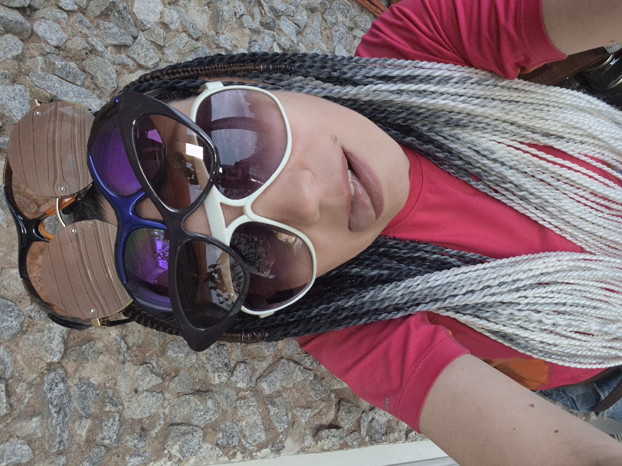 A person is lying down on a stone-tiled surface, angled so their head is towards the left of the frame and their feet towards the right. They are wearing a bright pink t-shirt. Their hair is styled in numerous thin braids, a mix of black, grey, and white strands. They are wearing multiple pairs of oversized sunglasses stacked on their face. The topmost pair has a large, thick white frame and brown tinted lenses. Below that, a pair of black-framed sunglasses with blue and purple mirrored lenses is visible. Another pair of sunglasses with brown tinted lenses and a black frame is positioned even lower. The person's mouth is slightly open, with pale lipstick applied. The background consists of the textured grey and brown stone tiles. The lighting suggests it is daytime, possibly sunny, as there are reflections in the sunglasses lenses. The overall impression is a casual, possibly outdoor setting. The image appears to be a selfie, taken from a high angle looking down at the person. There is no visible text.