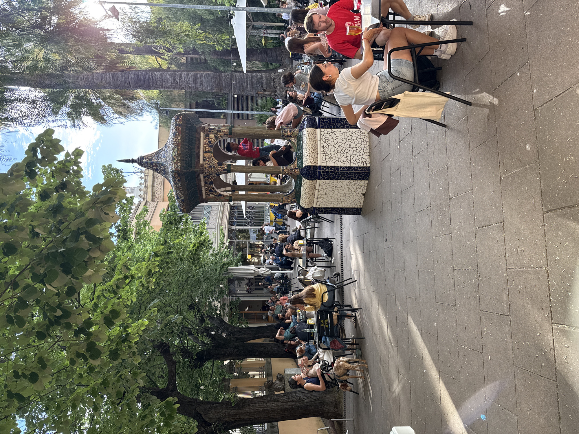 This image depicts a busy outdoor café scene in Barcelona, Spain. Many people are seated at tables, seemingly enjoying food and drinks. The area is shaded by trees and umbrellas, and there's a decorative, tiled fountain in the background. The setting appears to be a public square or pedestrian zone. There are numerous patrons, some conversing, others dining, and one person appears to be taking a photo. The environment seems relaxed and vibrant, typical of a Barcelona public space.