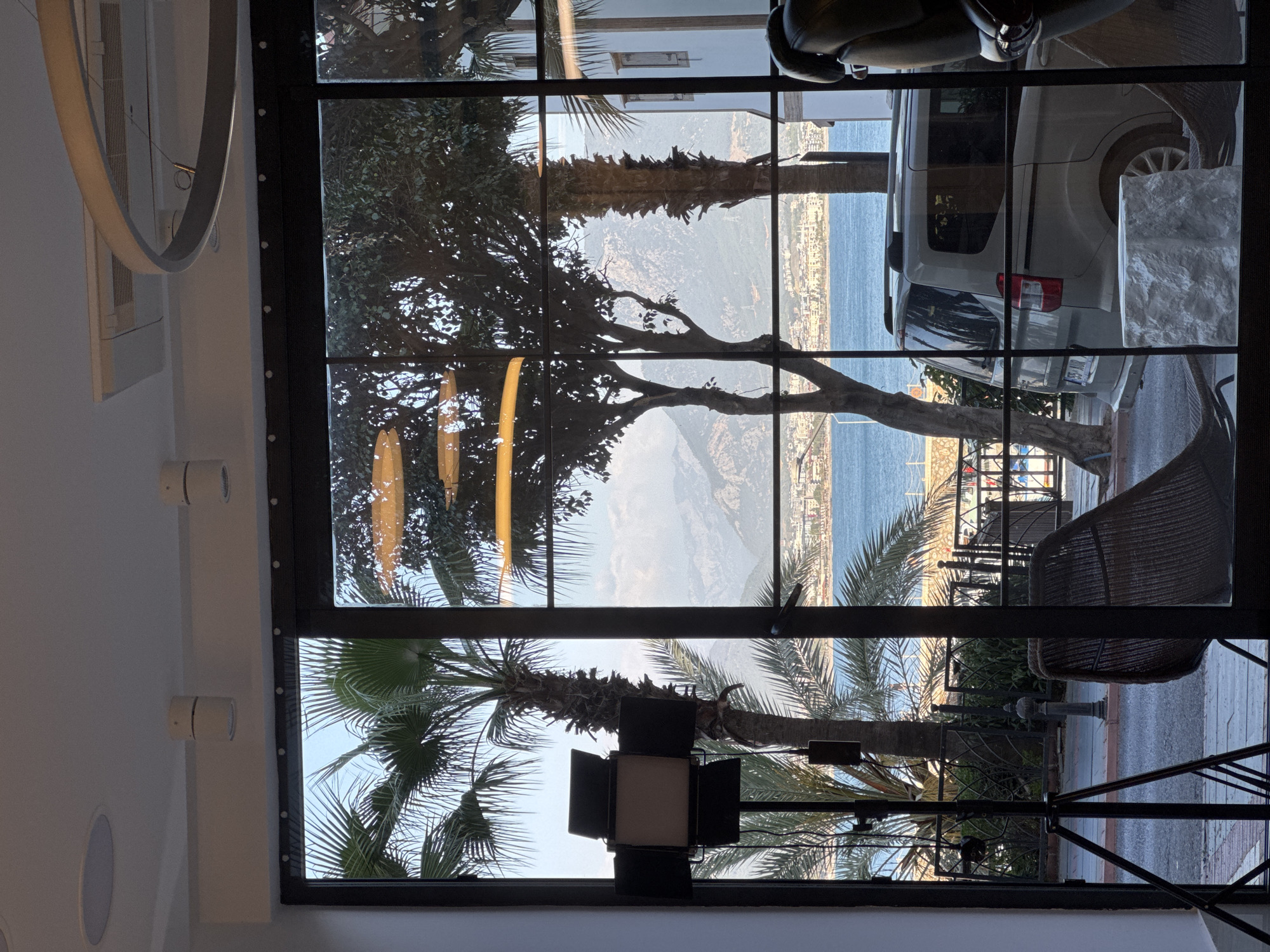 The image shows a view from inside a building in Alanya, Turkey, through a multi-paned window. The window frame is black. Outside, there is a view of the Mediterranean Sea, the Alanya coastline, a mountain, trees, and a white van parked on the street. Some chairs are visible on the street outside. Inside, a circular light fixture is seen on the ceiling.