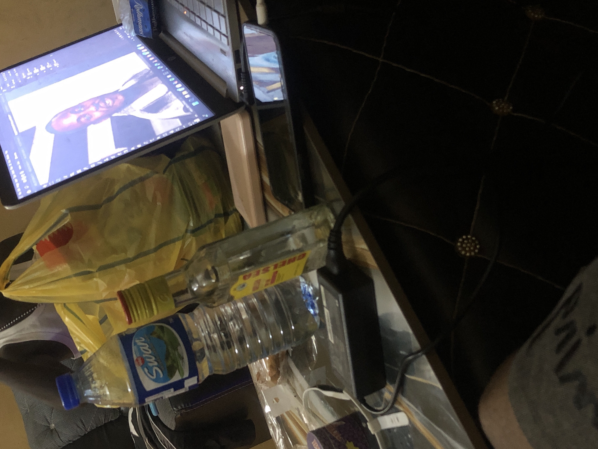 A person is sitting on a couch in Kabong, Nigeria, with a laptop open on a table in front of them. A bottle of Swan water and a bottle of alcohol are also on the table, along with a yellow plastic bag. The laptop screen displays a video call with another person. The room is dimly lit, and the floor has a diamond-patterned rug.