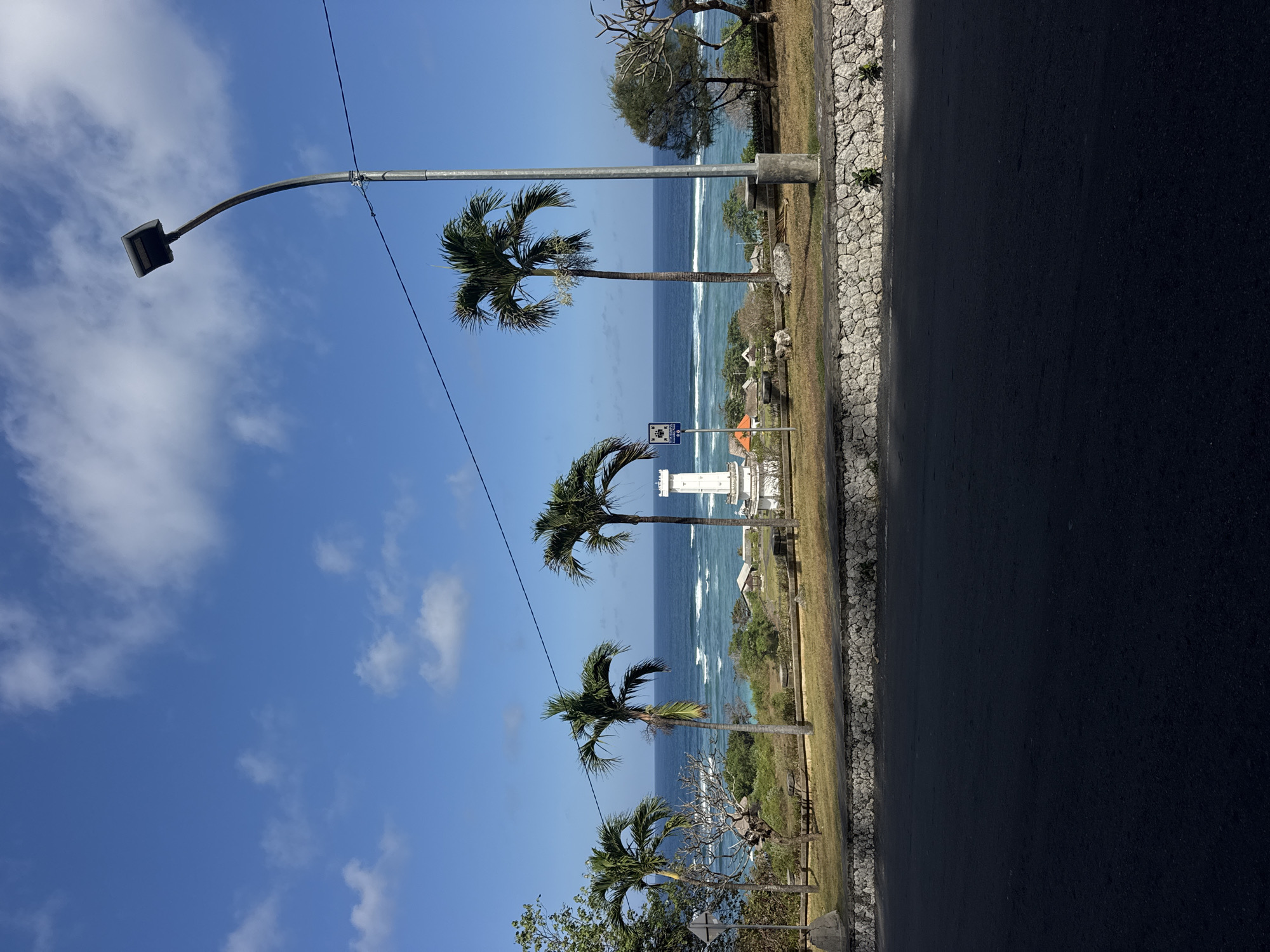 This is a landscape photograph taken in Kuta Selatan, Indonesia, featuring a coastal scene on a bright, sunny day with a clear blue sky and scattered white clouds. In the foreground, a dark asphalt road runs horizontally across the bottom of the frame, casting a shadow. Behind the road, a low, textured stone wall separates it from a grassy embankment.
Beyond the embankment, several palm trees are visible, with their fronds swaying gently. In the middle ground, the ocean stretches out, a deep blue with white-capped waves breaking near the shore. Along the coastline, there are several small buildings with orange roofs and some tropical vegetation.
A prominent white lighthouse or monument stands on the right side of the middle ground, with a dark blue sign depicting figures and text to its left. A modern street lamp with a dark fixture curves over the scene from the upper right. A thin, dark wire stretches horizontally across the upper part of the image. The overall impression is a serene, tropical coastal view.
