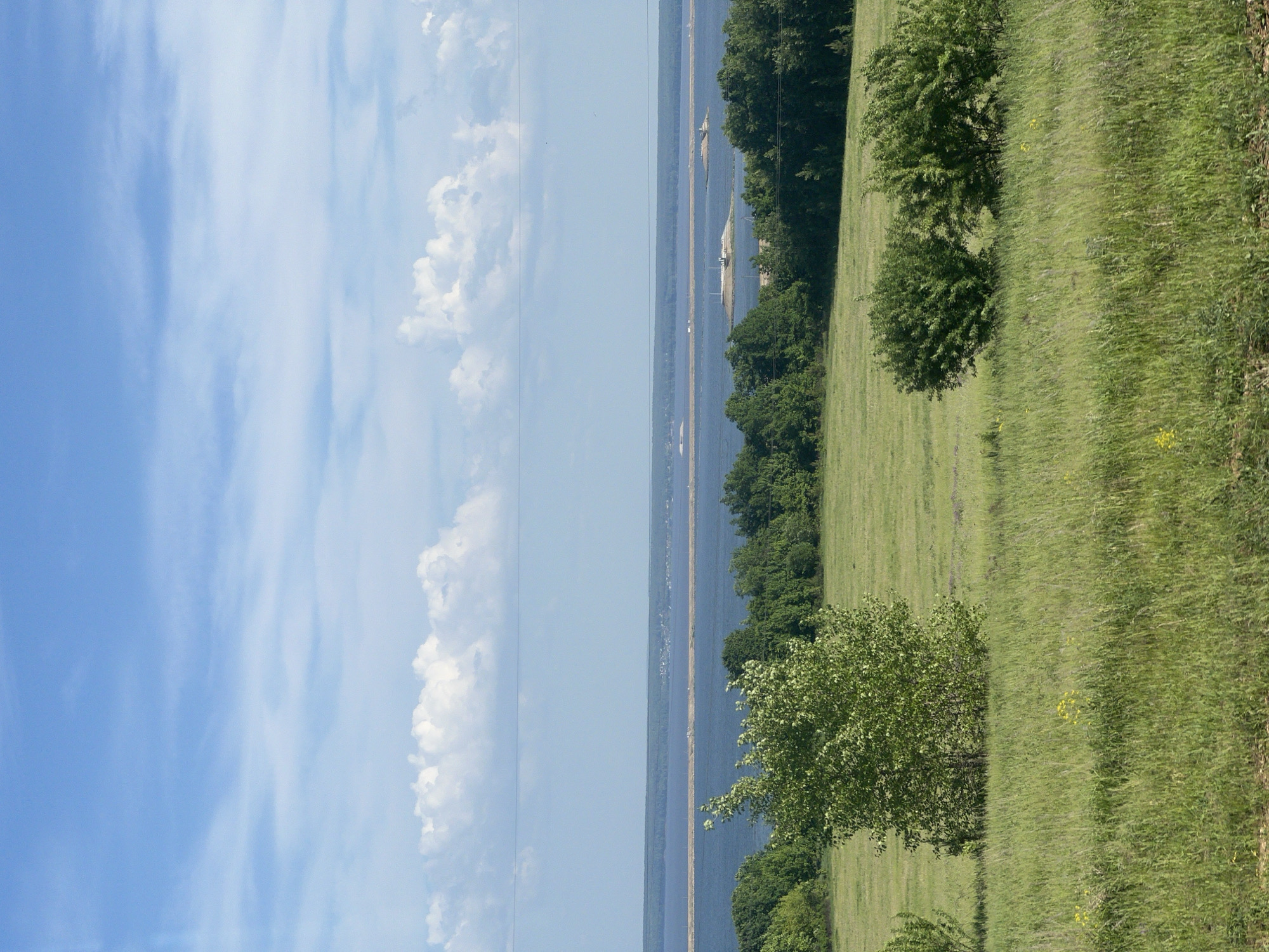 The image presents a landscape view of Белоус, Russia. The sky, with a mix of blue and white clouds, covers the left portion of the frame. Adjacent to the sky, a body of water, likely a river or a lake, extends horizontally across the middle, bordered by a thin land strip in the distance. The right side of the image depicts a grassy field dotted with trees and bushes. The field has varying shades of green, suggesting different types or conditions of vegetation.