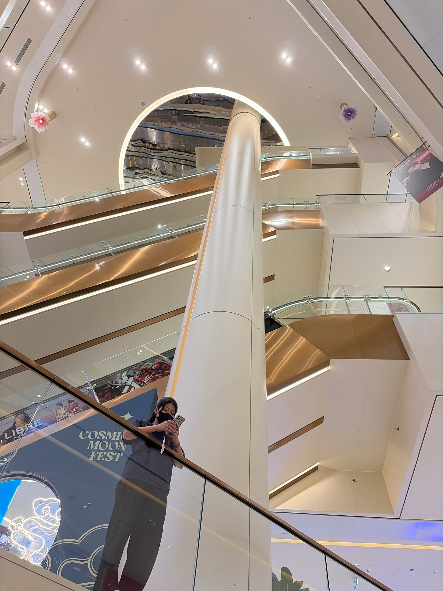 The image shows the interior of a modern shopping mall with multiple levels and escalators. A person is standing on an escalator, looking at their phone. The mall features a large central column, geometric architecture, and decorative lighting. In the background, there are posters and displays for various brands and events. The scene appears to be bright and spacious, with clean lines and a contemporary design. The presence of a person using a phone suggests a normal activity within a public space. The overall environment indicates a commercial setting designed for shopping and entertainment. The text visible on a display in the lower left corner, "COSMIC MOON FEST," suggests a seasonal event or promotion.