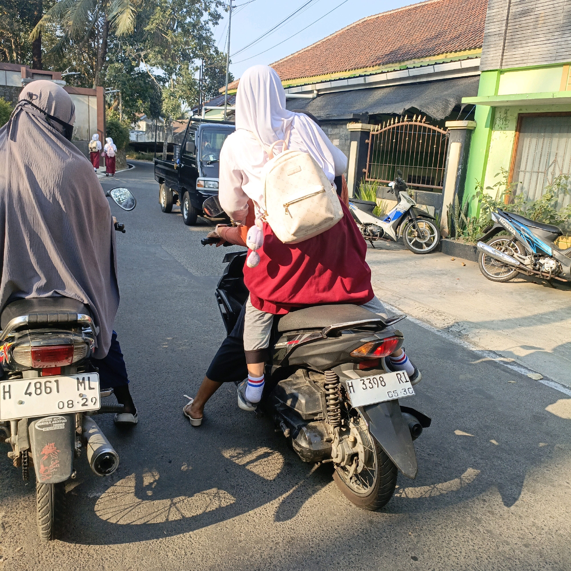 In Salatiga, Indonesia, two young women wearing hijabs and school uniforms are riding on a motorcycle, with one in the front driving and the other as a passenger behind her. The passenger wears a white hijab, a white shirt, and a red skirt, carrying a cream-colored backpack adorned with a bunny keychain. The driver wears a grey hijab and a dark-colored outfit. They are traveling down a paved road lined with houses, fences, and parked motorcycles. A black truck is visible in the background, and the sunlight casts distinct shadows of the motorcycles and a bicycle wheel on the road surface. The license plates visible on the motorcycles read "H 4861 ML 08.29" and "H 3398 RI 05.30". The scene appears to be a typical street view in the city, possibly during school hours given the attire of the individuals.