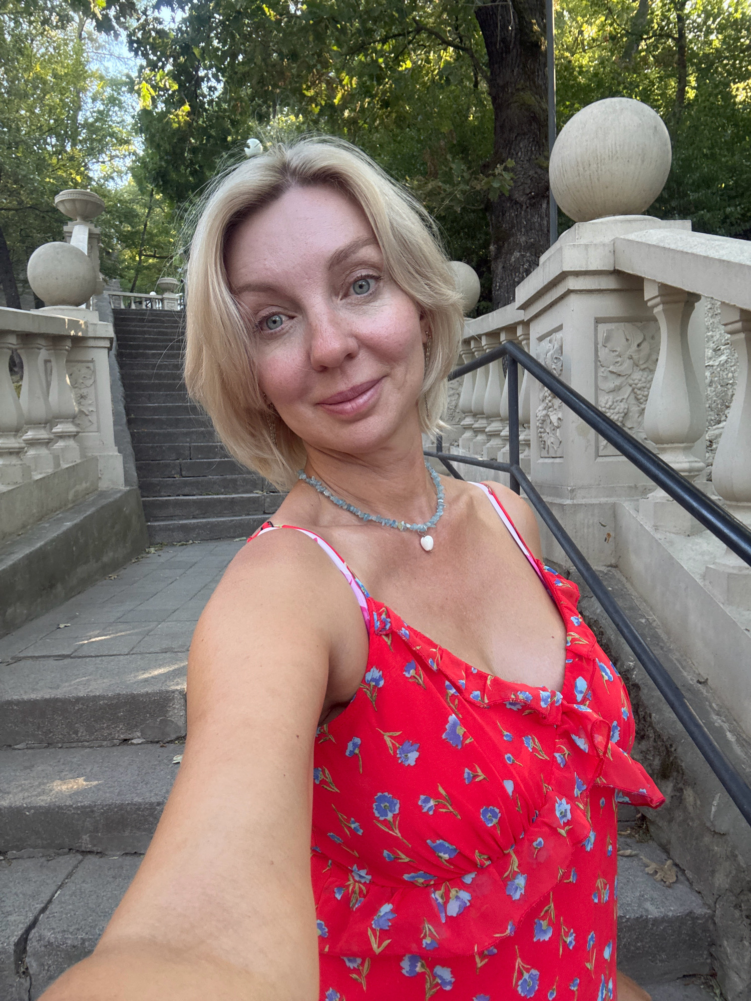 In Abrau-Dyurso, Russia, a woman with blonde hair is taking a selfie on a stone staircase outdoors. She is wearing a red dress with a floral pattern and a necklace. The staircase is lined with ornate stone railings and balustrades, and mature trees with green foliage are visible in the background. The lighting suggests it is daytime, likely morning or late afternoon, with the sun casting shadows on the steps.