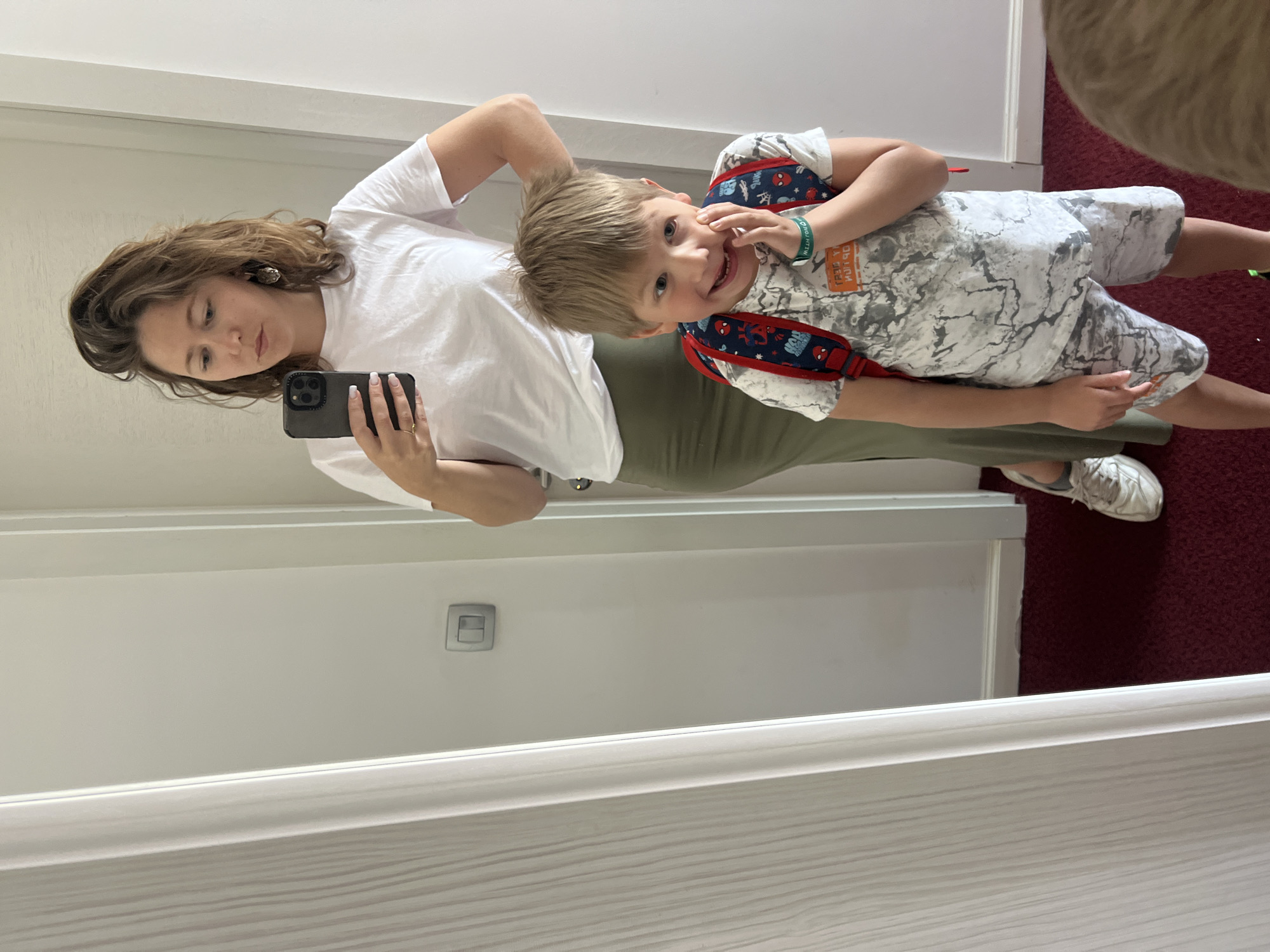 The image, taken in Petrovac centar, Montenegro, captures a mirror selfie of a woman and a young boy indoors. The setting appears to be a well-lit room or hallway, featuring a large mirror against a white wall, a white baseboard, and a dark red carpet.
The woman, with shoulder-length brown hair, is positioned to the left, wearing a white short-sleeved t-shirt, olive green pants, and white sneakers. She is holding a dark-colored smartphone to take the picture, with her nails painted white and a ring visible on her finger. She wears earrings and looks towards the phone with a composed expression.
To her right, slightly in front, stands a cheerful young boy. He is smiling widely at the camera, with light brown hair and bright eyes. He wears a white t-shirt with a gray and white marble-like pattern and what appear to be matching shorts. A red and blue Spider-Man themed backpack is on his back, with the straps visible over his shoulders. On his left wrist, he wears a green silicone wristband.
Visible text includes inverted characters on the boy's t-shirt, which partially reads "KEEP YOUR EYES OPEN," and on his wristband, which reads "I LOVE PETROVAC." The Spider-Man backpack also features small Spider-Man logos and text. The bright lighting indicates it is likely daytime, and the overall atmosphere suggests a casual, happy moment, possibly before an outing, given the boy's backpack.