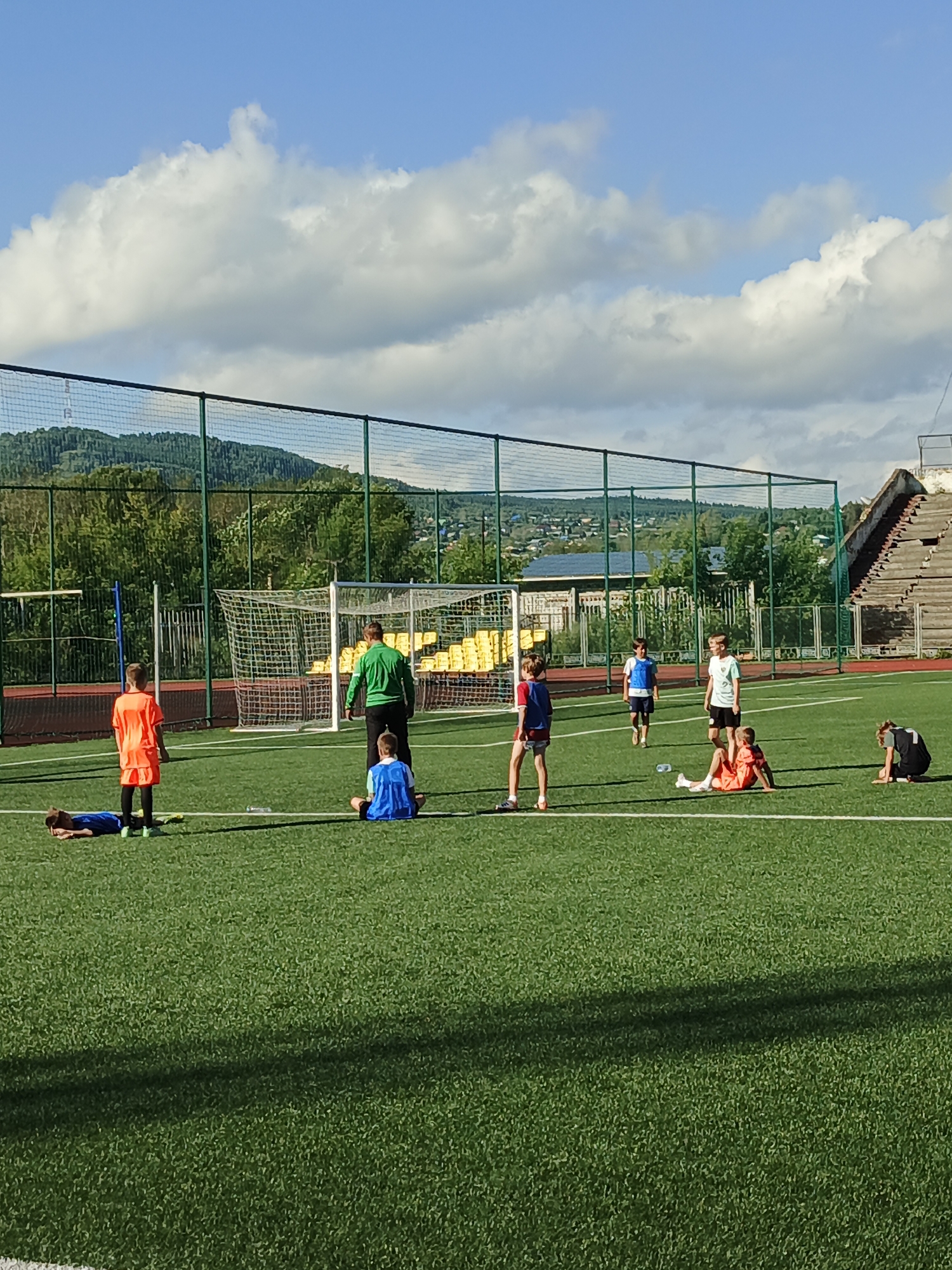 The image depicts an outdoor soccer training session for children on an artificial turf field, situated in Zlatoust, Russia. A group of approximately eight children and one adult are visible on the field. The adult, dressed in a green top and dark pants, appears to be coaching or supervising. Children are in various states of activity, with some sitting on the ground, one lying down, and others standing. They are wearing athletic attire, primarily t-shirts and shorts, in vibrant colors like orange, blue, and white.
The training field is marked with white lines and features a soccer goal in the background. Beyond the field, a fence encloses the area, and behind that, a dense line of green trees and a hillside with scattered buildings are visible, suggesting a semi-urban or suburban setting within Zlatoust. A tiered stadium structure, possibly disused, is partially visible on the right side of the image. The sky above is blue with scattered white clouds, indicating fair weather. The overall scene conveys a sense of organized sports activity taking place in a natural, yet developed, environment.