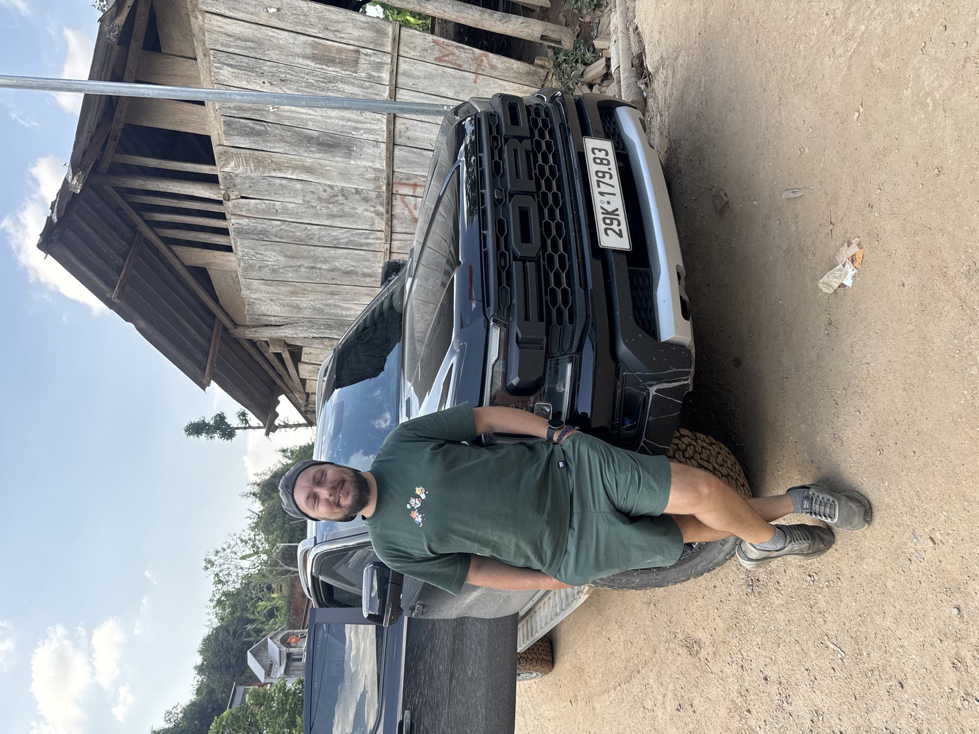 The scene depicts a man standing next to a dark Ford pickup truck in Pờ Ly Ngài Commune, Vietnam. The truck has a license plate number of "29K-179.83". The man is wearing a green t-shirt with designs, matching shorts, a grey beanie, and grey sneakers. He's leaning against the front of the truck. Behind the truck is a weathered wooden structure. The ground is dirt. The sky is visible with scattered clouds.