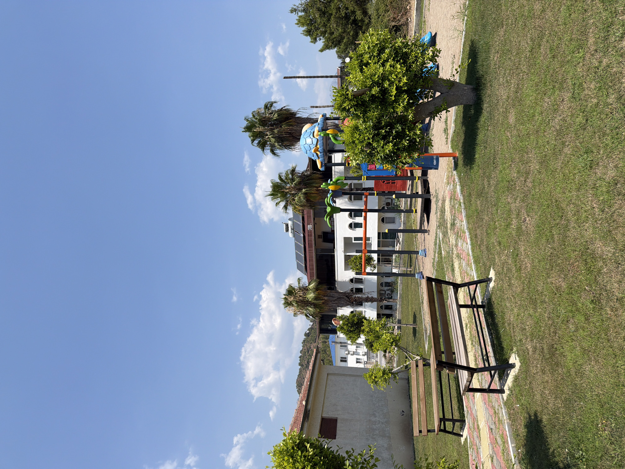The image depicts a children's playground located in Kemer, Turkey, under a clear blue sky with scattered clouds. In the foreground, two wooden benches with black metal frames are positioned on a patch of green grass. Behind the benches, a pathway made of interlocking bricks and concrete borders a grassy area.
The playground features a colorful play structure with a blue turtle-shaped canopy. The structure includes various colorful plastic elements in red, yellow, and blue, along with metal bars for climbing and swinging. Adjacent to the play structure, a small tree with vibrant green foliage is situated. In the background, several buildings are visible. To the left, a low-rise building with a tiled roof and beige walls stands. To the right, a multi-story white building with balconies and arched windows is present, topped with solar panels. Palm trees are scattered throughout the scene, adding to the Mediterranean atmosphere. The ground of the playground area appears to be a mixture of sand and grass. A tall, slender pole stands to the right of the play structure.