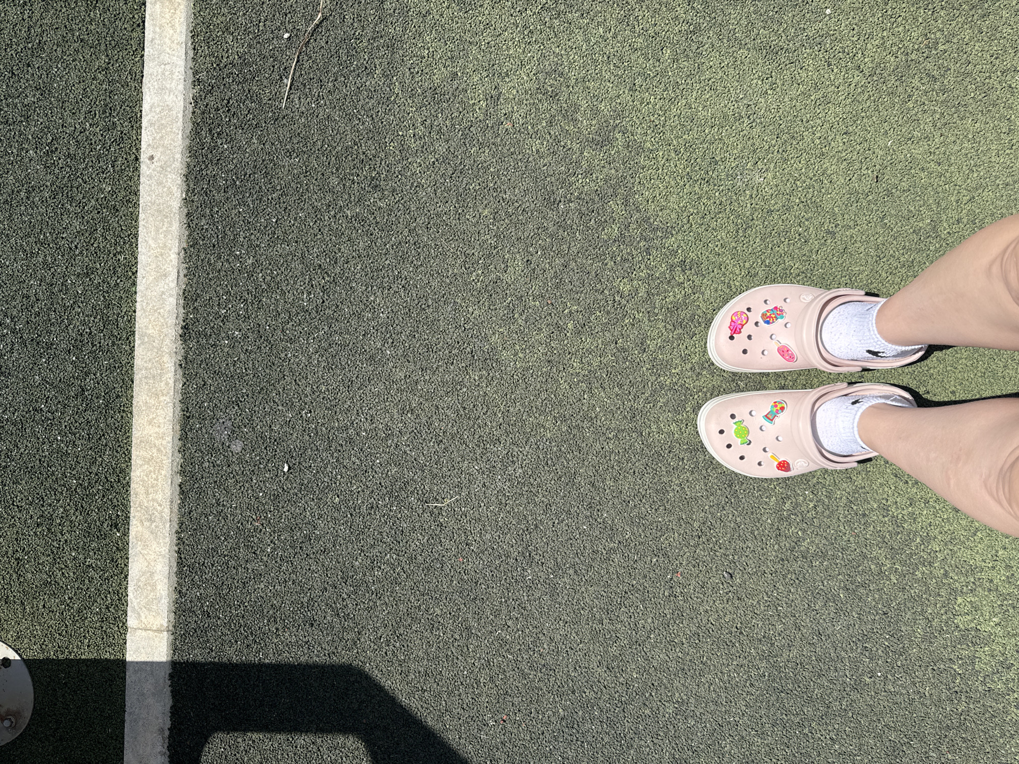 In Vyazniki, Russia, on a sunlit day, a pair of light pink Crocs, adorned with colorful Jibbitz charms resembling candy and fruit, are worn by an individual standing on a dark green, granular surface. The shoes are paired with white socks. A thin, light-colored line runs horizontally across the upper portion of the image, marking a boundary on the textured ground. A shadow cast by an unseen object falls diagonally across the upper left quadrant of the scene. The overall environment suggests an outdoor recreational area or playground.