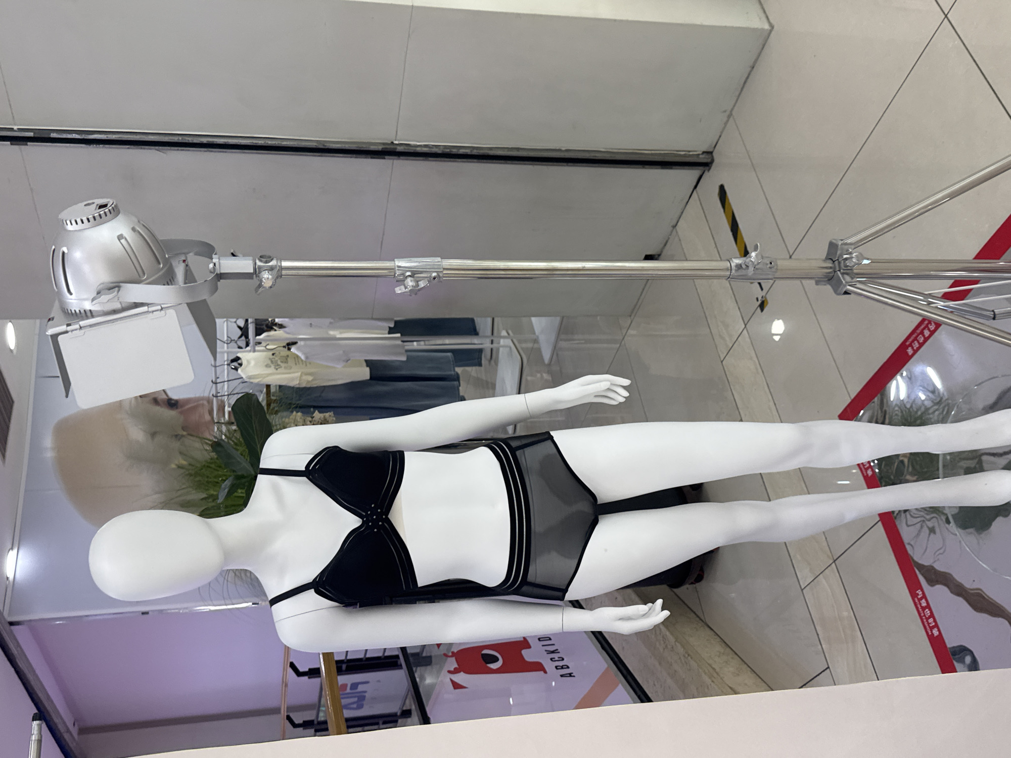 The image depicts a white female mannequin in a retail setting, possibly a clothing store. The mannequin is positioned centrally and is wearing a black bra and matching sheer black underwear. To the right of the mannequin, a tall, silver lighting stand with a rectangular light fixture is visible. In the background, racks of clothing, including shirts and pants, can be seen. A large advertisement featuring a stylized red monster is on a wall to the left of the mannequin. The floor appears to be tiled, with some red safety markings and reflective material on the floor in the foreground. The overall scene suggests a retail display or a photo setup within a store in Linghe, China.