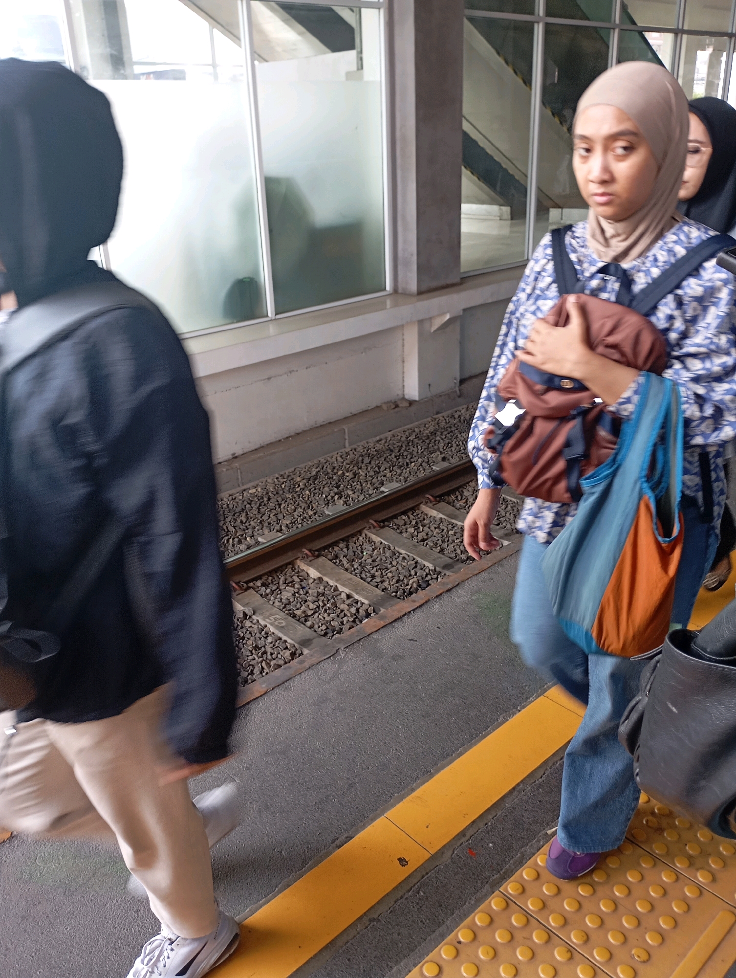 Two individuals are on a train platform in West Jakarta, Indonesia, likely near a station. The person in the foreground is wearing a black hijab, a black jacket, and light-colored pants, with one white sneaker visible. They are walking away from the camera, with their back and left side partially visible. The second individual, to the right and slightly behind, is wearing a light brown hijab and a blue and white patterned shirt, along with blue jeans. They are carrying a brown backpack and a blue and orange tote bag. The background shows train tracks with gravel and wooden ties, a concrete platform edge with a yellow safety line, and a modern building with large glass windows. The lighting suggests it is daytime.