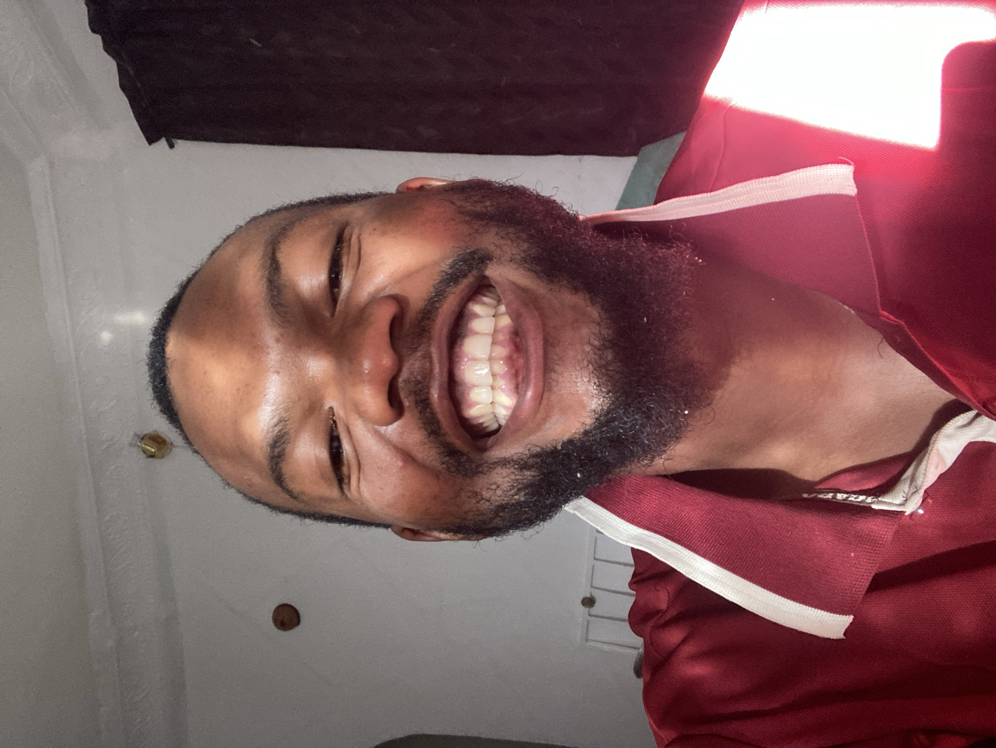 A man with dark skin and a full beard is smiling widely, exposing his teeth. He is wearing a red shirt with white trim at the collar. The background includes a light gray wall with a few small objects attached, and a dark colored fabric hanging near the top of the frame. There is strong light source above, to the right of the man, creating a bright area on the wall and causing red color distortion on the right side of the man's shirt. The location is Jos, Nigeria.
