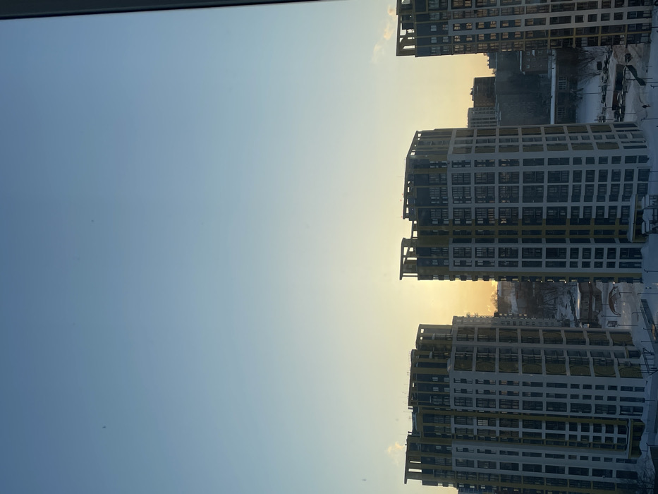 The image depicts an urban landscape in Izhevsk, Russia, during winter, captured from an elevated perspective, likely a residential window. The scene is illuminated by a low-angle sun, indicating either sunrise or sunset, with a sky transitioning from light blue overhead to a bright yellow-orange at the horizon.
In the mid-ground, two tall, modern residential buildings with distinctive architecture dominate the frame. These multi-story structures feature facades composed of light-colored panels accented by prominent vertical and horizontal yellow-gold banding, with numerous rectangular windows. The sun's intense light emanates from behind and between these two buildings, casting their inner facades into silhouette.
The ground is uniformly covered in a thick layer of white snow, extending from the immediate foreground into the distant background. Visible urban elements include smaller, less distinct residential buildings, snow-covered roads, and parked vehicles, some also blanketed in snow. Several small, dark figures, identified as pedestrians, are discernible on snow-cleared pathways below the prominent buildings. Bare deciduous trees are scattered throughout the snowy landscape.
The architectural style of the prominent buildings, particularly the white and yellow-gold paneling, is consistent with the "Volna" residential complex located in Izhevsk, Russia, such as those found near Udmurtskaya Street.