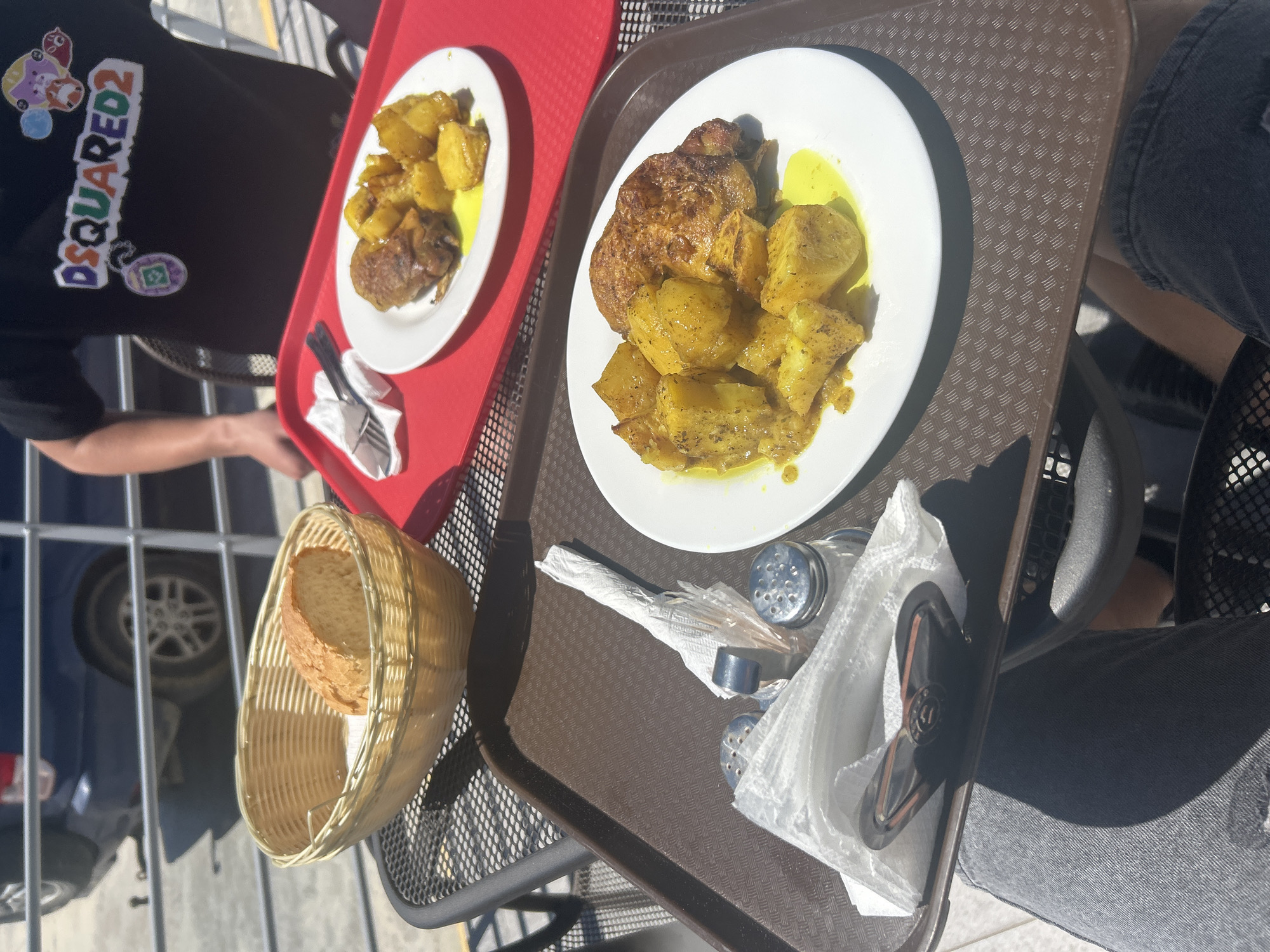 The image shows a meal setting in what appears to be an outdoor dining area in the city of Νέα Κερδύλια, Greece. There are two plates of food containing what appears to be chicken and potatoes with a yellow sauce. A basket of bread is present along with utensils, salt and pepper shakers and napkins. Two individuals are visible. One is wearing a black T-shirt with "DSQUARED2" written on it and another person wearing gray denim jeans. The food items are on two separate trays. The location appears to be outdoors with visible parts of a car's tire.