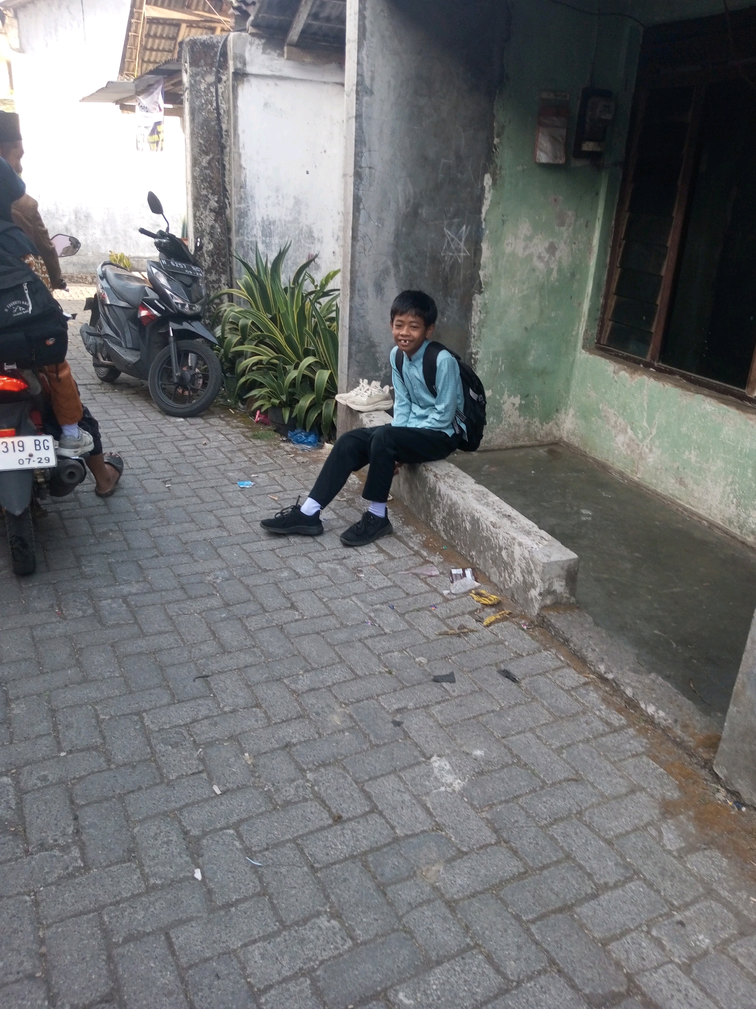 A young boy with short black hair and a light blue shirt with black pants and a black backpack is sitting on a concrete ledge outside a building. He is smiling and looking towards the camera. Next to him, a motorcycle is parked on a brick-paved walkway. In the background, there is a building with a green, weathered exterior and a window. The area appears to be an urban or semi-urban environment in Andongsili, Indonesia, with a house or shopfront visible. The walkway has some debris scattered on it. Another person is partially visible on a motorcycle to the left of the frame.