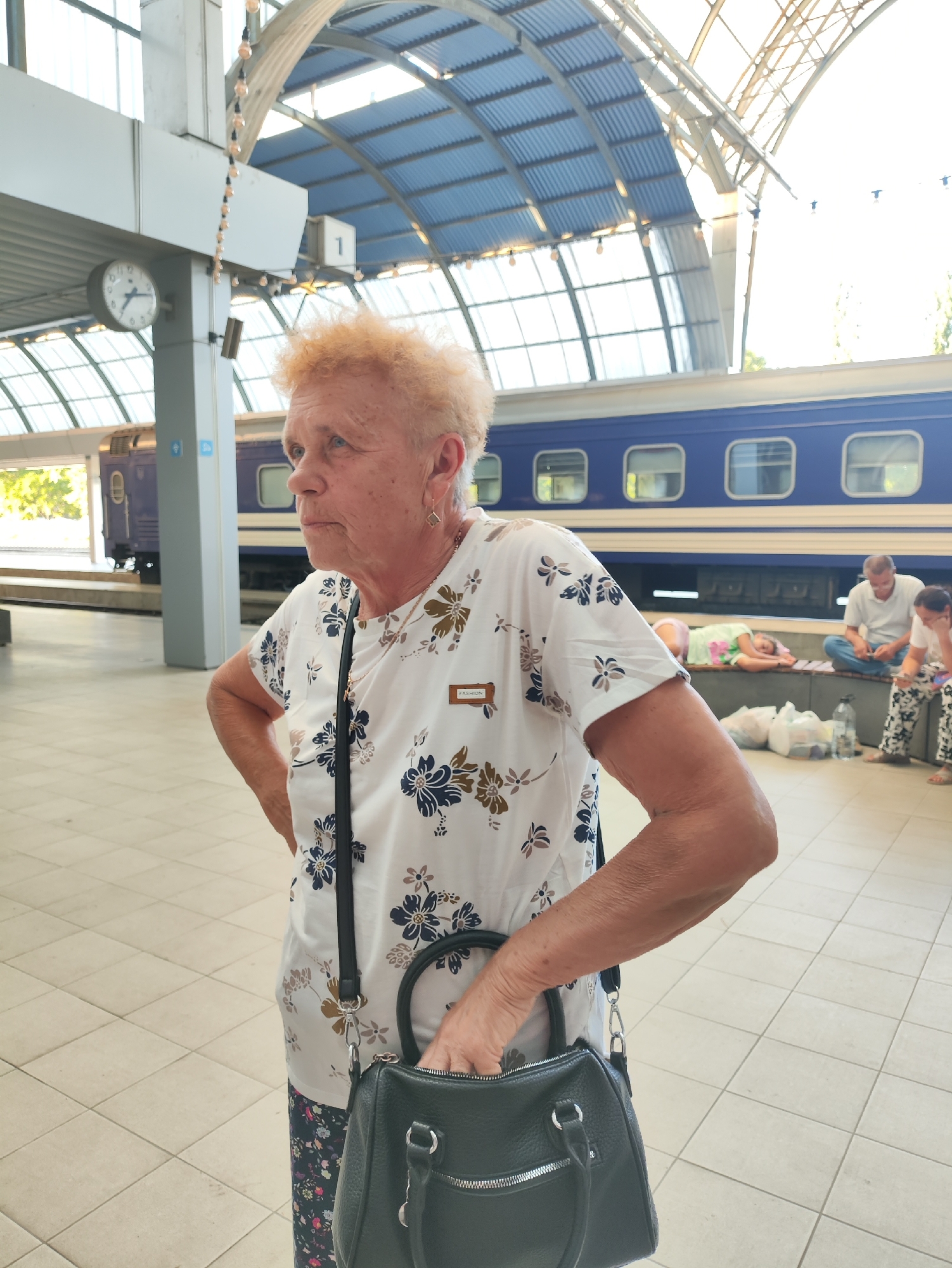 The image depicts an elderly woman with orange hair standing in what appears to be a train station waiting area. She is wearing a white t-shirt with a floral pattern and dark pants, and she is holding a black handbag. In the background, a blue train is visible, and other people are seated on a bench. The setting features a tiled floor and a covered roof structure with visible supports and lights. The overall scene suggests a moment of waiting or transit in a public transportation hub.