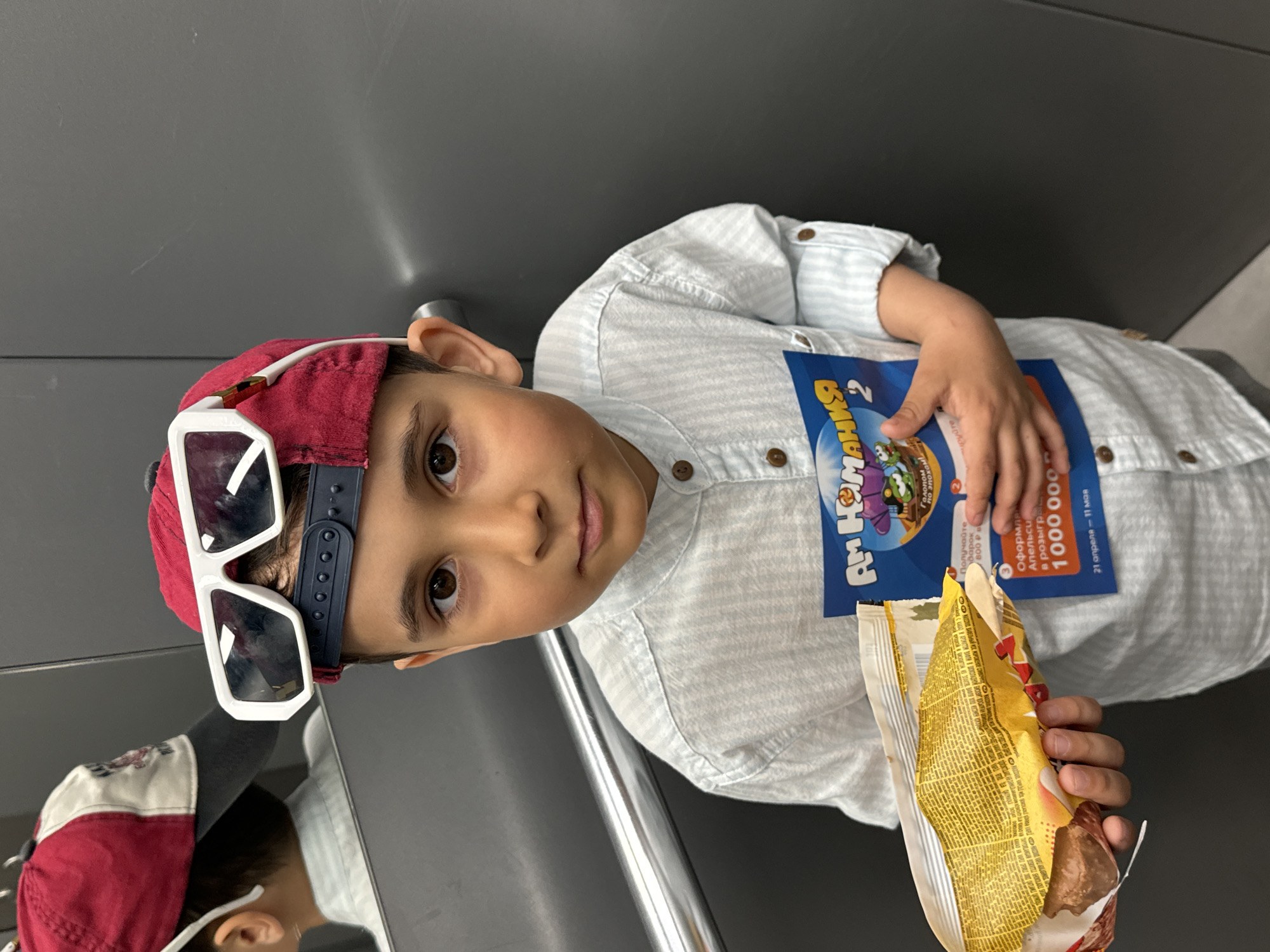 A boy is posing indoors, likely in a public space like a restaurant or transit station, in Chaikovsky, Russia. He is wearing a red baseball cap with a pair of white sunglasses perched on the brim. He is holding a partially opened snack package in his right hand and a blue advertisement brochure in his left hand. The brochure is titled "Ам Кумания 2" and mentions receiving a gift of 800 rubles. It also mentions a raffle or lottery with a prize of 1,000,000, taking place from April 21 to May 11. The background is a gray wall or panel. There is a reflection of the boy and his hat visible on the lower left.