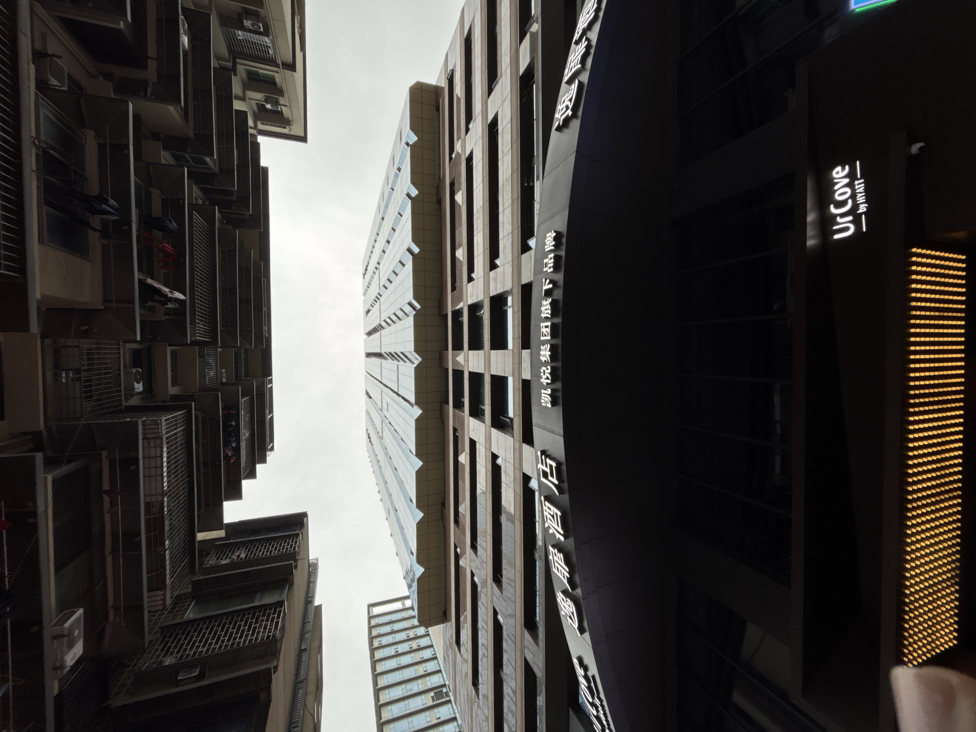 The image shows a view looking straight up at buildings in Yuzhong District, China. On the left are older residential buildings with external air conditioning units and metal-framed balconies, some with visible clotheslines. In the center are taller modern buildings, one with a reflective glass facade. On the right, a dark building displays the sign "UrCove - by HYATT" and vertical Chinese text, with a column of lights. The sky is overcast.