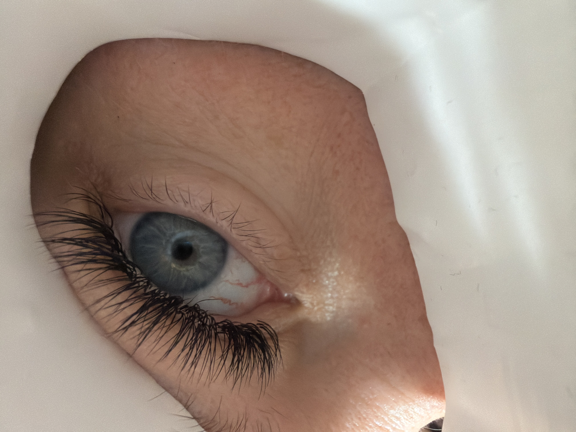 A close-up image shows a human eye peeking through a white material, possibly fabric or paper. The eye has a blue iris and long, dark eyelashes. Some veins are visible in the white of the eye. The skin surrounding the eye is light-toned. The image is taken in a well-lit environment. No information in the image indicates a location in Chaikovsky, Russia.
