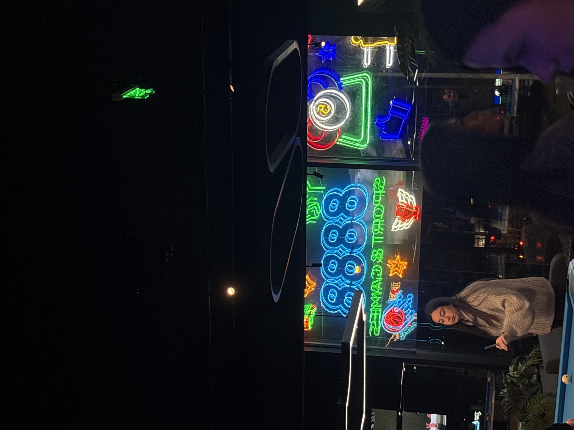The image shows a nightlife scene, likely a bar or club in Varna, Bulgaria. The main focus is a woman in a beige sweater and dark pants playing pool. There are neon signs with various shapes and colors visible, including stars, horseshoes, and Chinese characters, as well as an arcade sign. A neon green pool table image is visible on the left side of the picture. Other patrons are blurred in the background, suggesting a crowded environment. The overall lighting is dim, with neon lights providing the primary illumination.