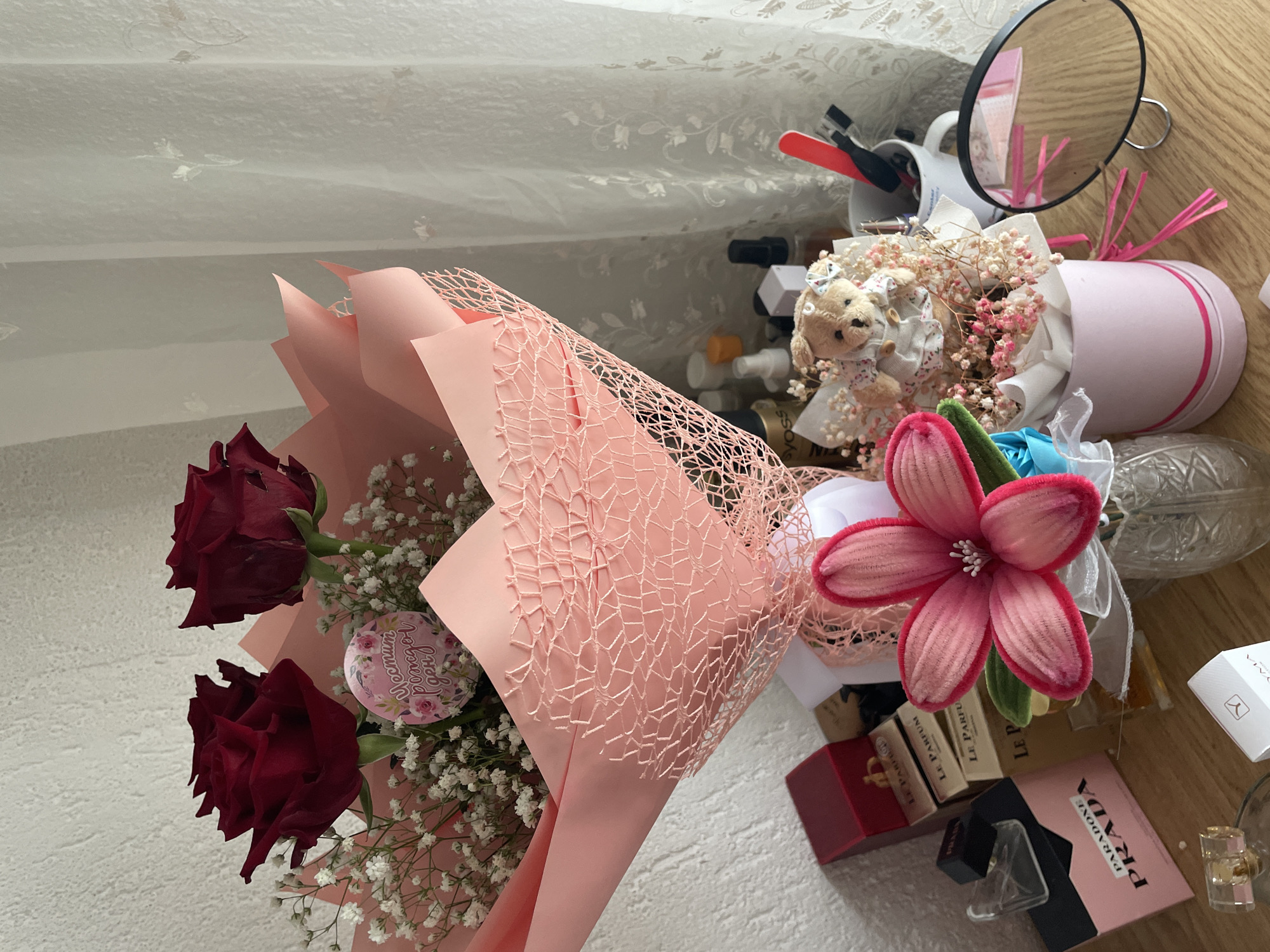 The image presents a cluttered tableau on a wooden surface, dominated by two floral arrangements. One bouquet features two dark red roses accompanied by delicate white baby's breath, wrapped in pink paper and a pink netting. Attached to the bouquet is a small, round sticker bearing a floral design. Adjacent to this bouquet is another arrangement, a pink cylindrical container filled with a teddy bear wearing a frilly dress and surrounded by dried flowers. A pink artificial flower lies near the base of this arrangement.
Various items surround these focal points, including a selection of miniature perfume bottles and boxes from brands such as Prada and Le Parfum. A magnifying mirror, a white mug filled with sundry items, and a decorative glass bottle are also visible. The background features a light-colored curtain with a subtle pattern.
The location in the city of Zlatograd, Bulgaria, cannot be determined from the image alone.